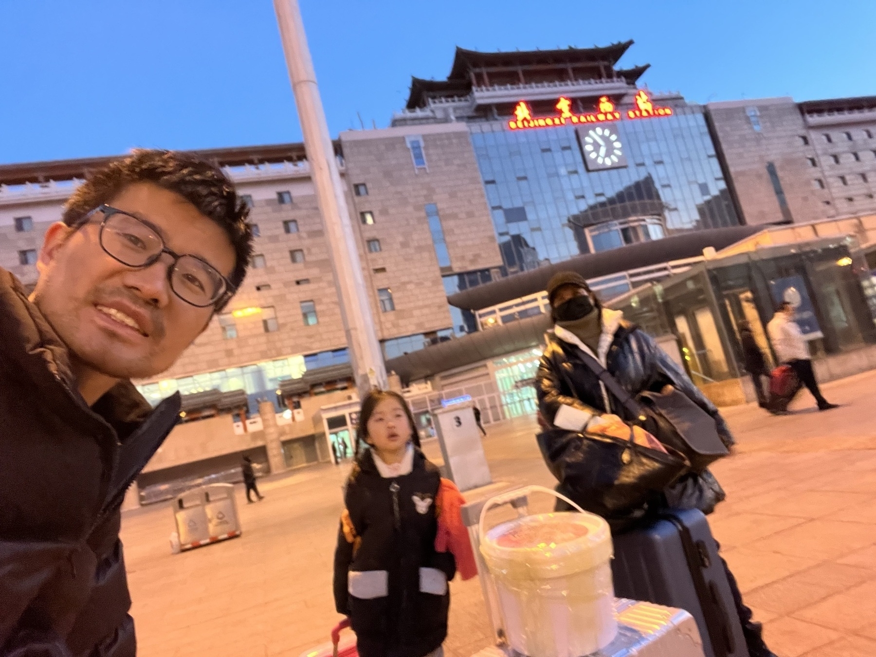 A man takes a selfie in front of a large building with a clock, accompanied by a child and another person with luggage.