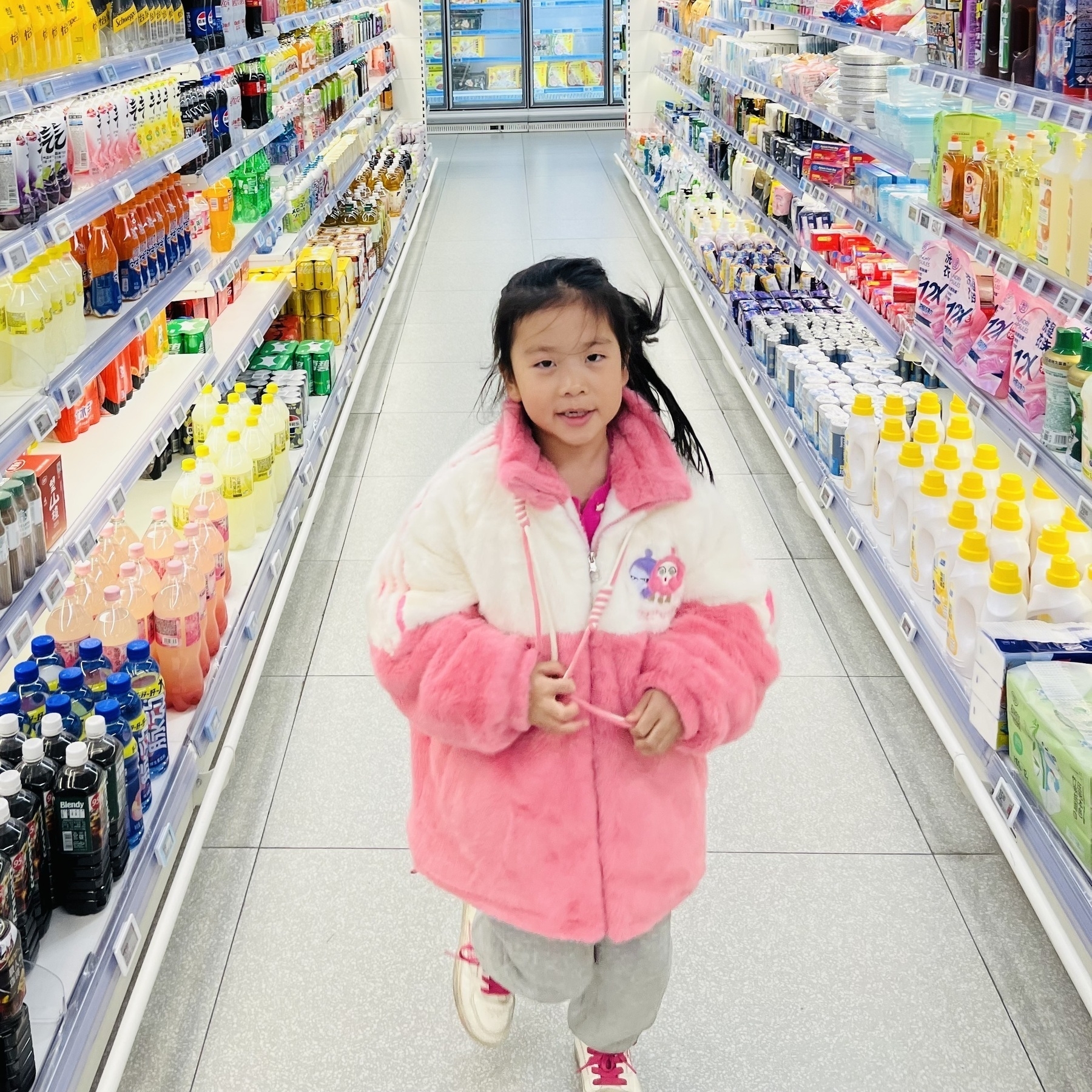 A young girl wearing a pink and white coat walks down an aisle in a supermarket filled with various beverages and household items.