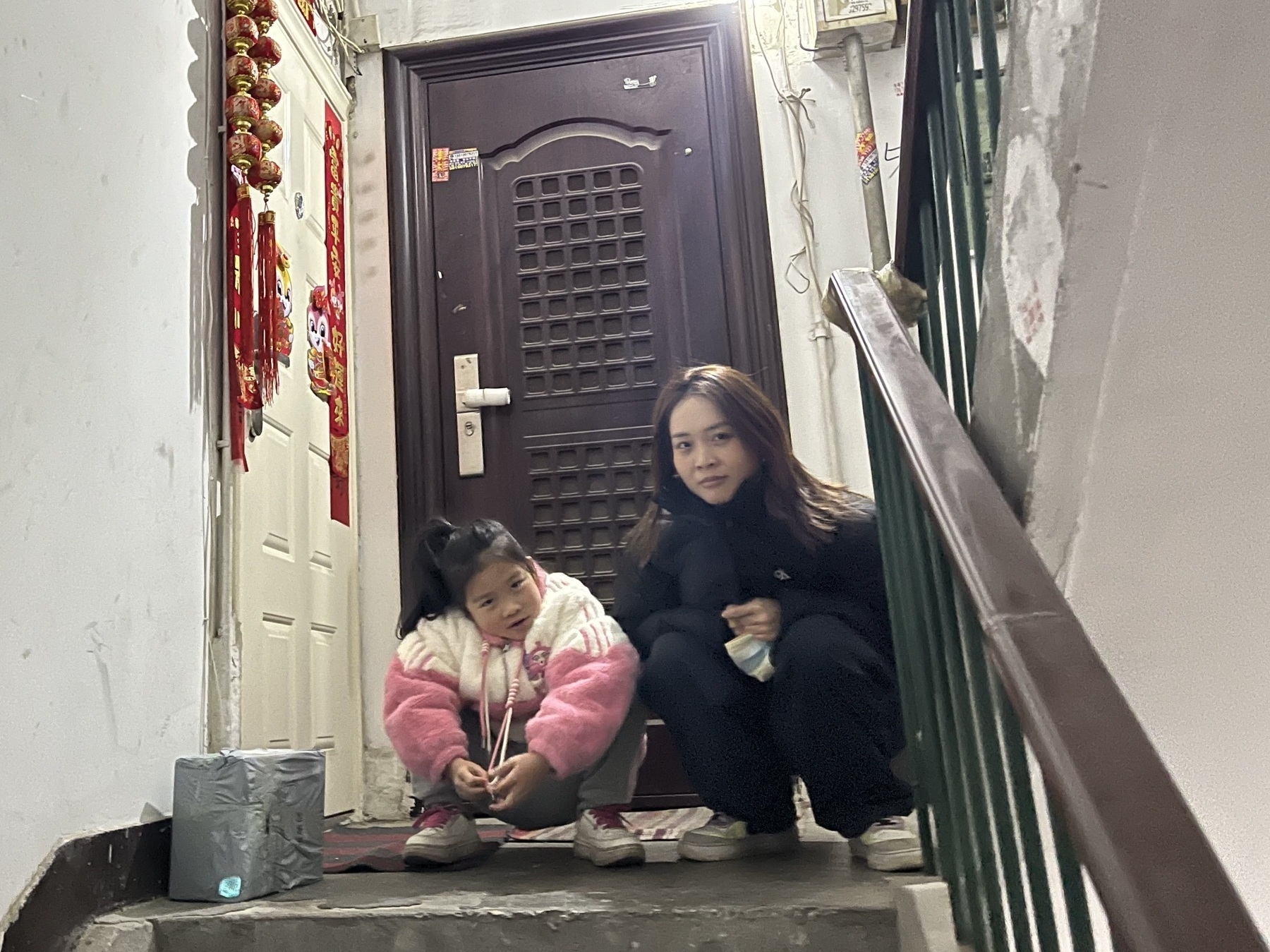 Two people are seated on a stairway landing in front of a door, with one holding a tissue box and decorative items hanging nearby.