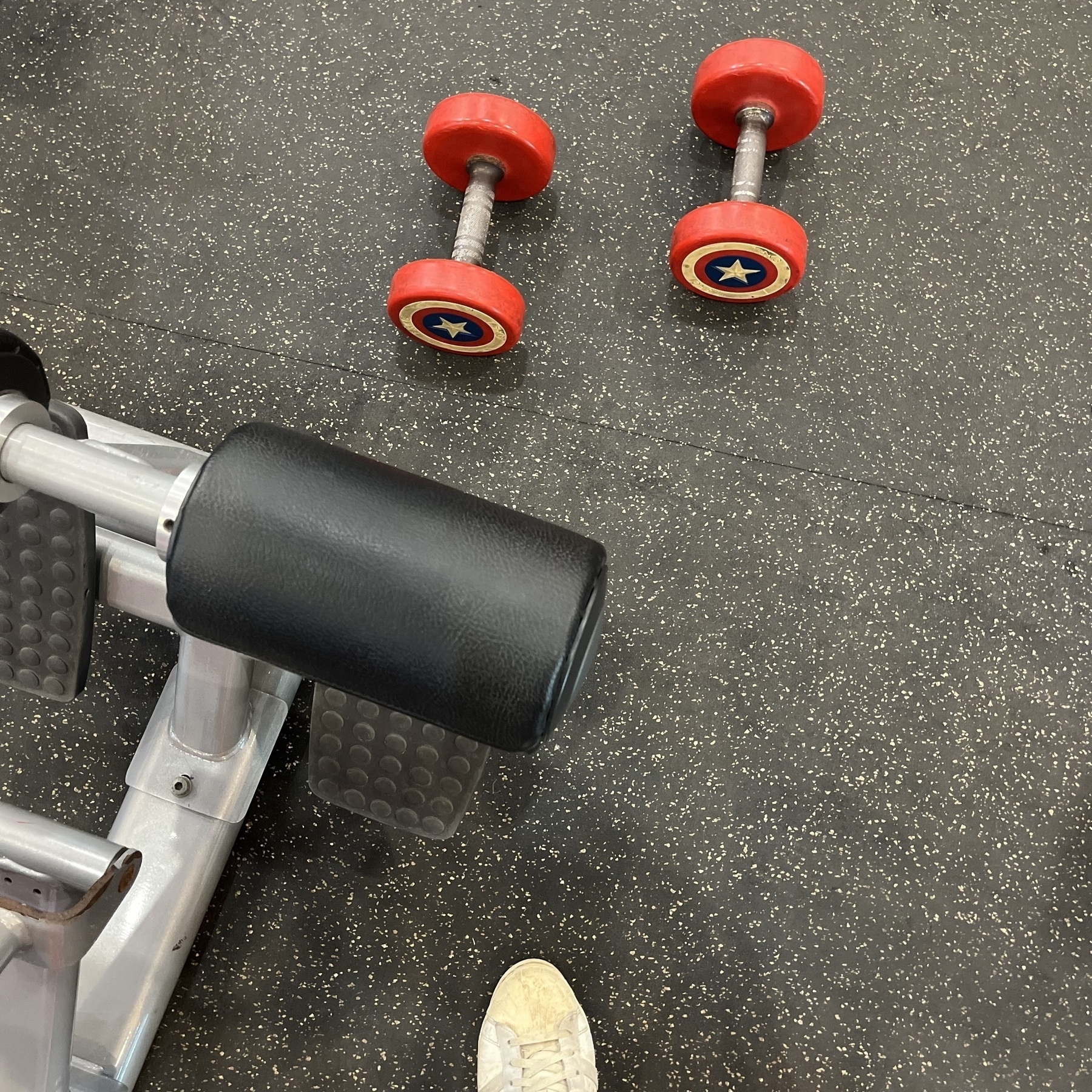 A gym setting includes two dumbbells with red weights and a black padded exercise machine, with a partial view of a person's shoe.