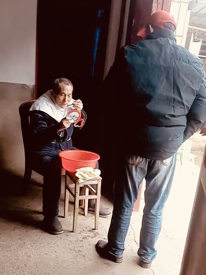 Uncle is shaving. A person is sitting and holding a bowl while another stands nearby in what appears to be an indoor setting.