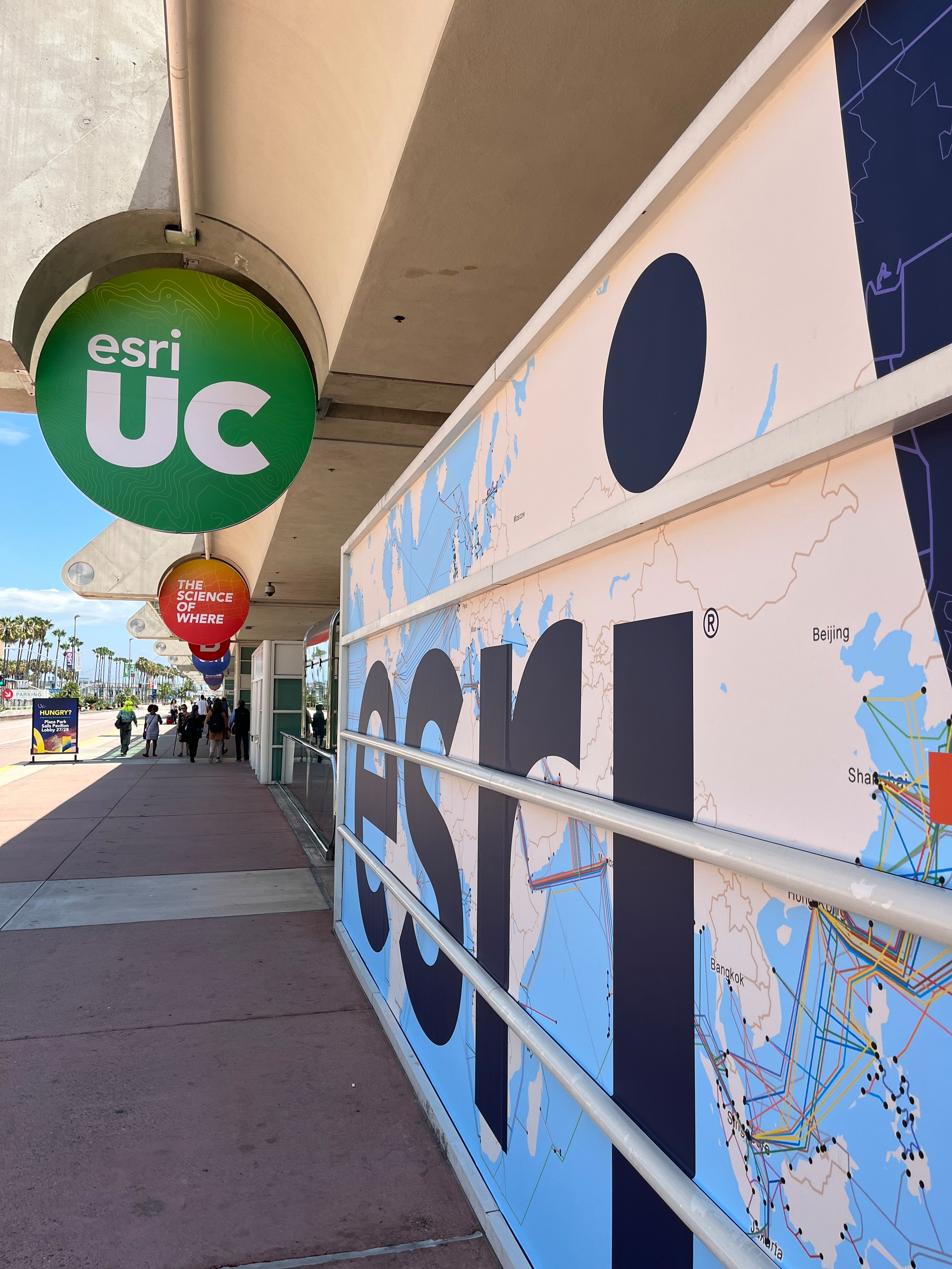 A sidewalk next to a large wall display features the words esri and maps, with circular signs hanging above that read esri UC and THE SCIENCE OF WHERE.