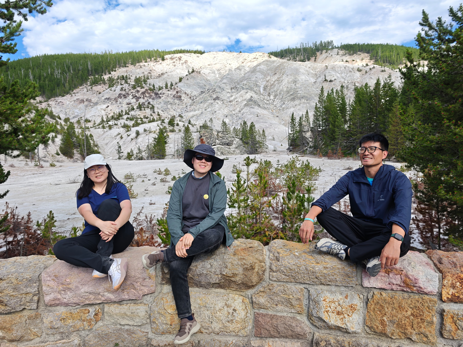 Three people are sitting on a stone wall with a scenic view of rocky hills and pine trees in the background.