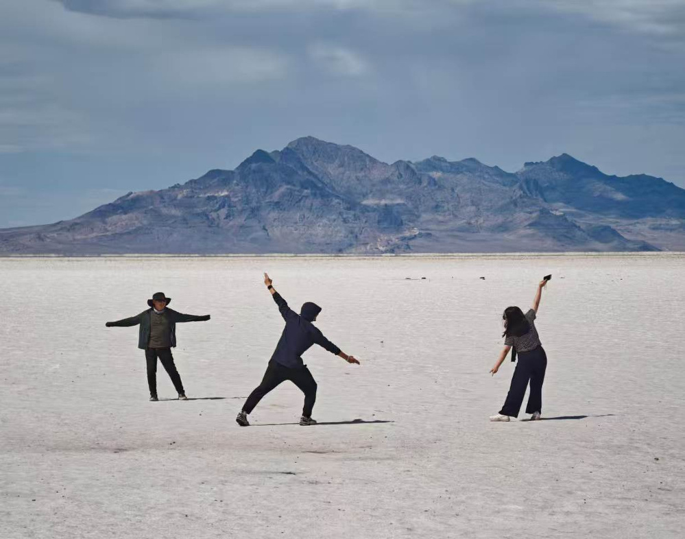 Three people are striking playful poses on a vast, flat salt plain with a mountain range in the background under a cloudy sky.