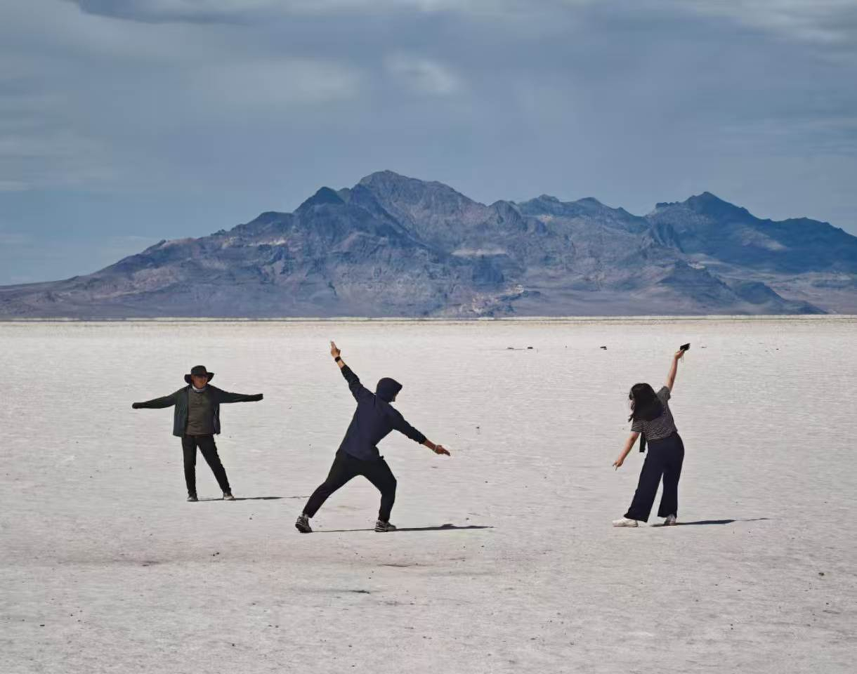 Three people are striking playful poses on a vast, flat salt plain with a mountain range in the background under a cloudy sky.