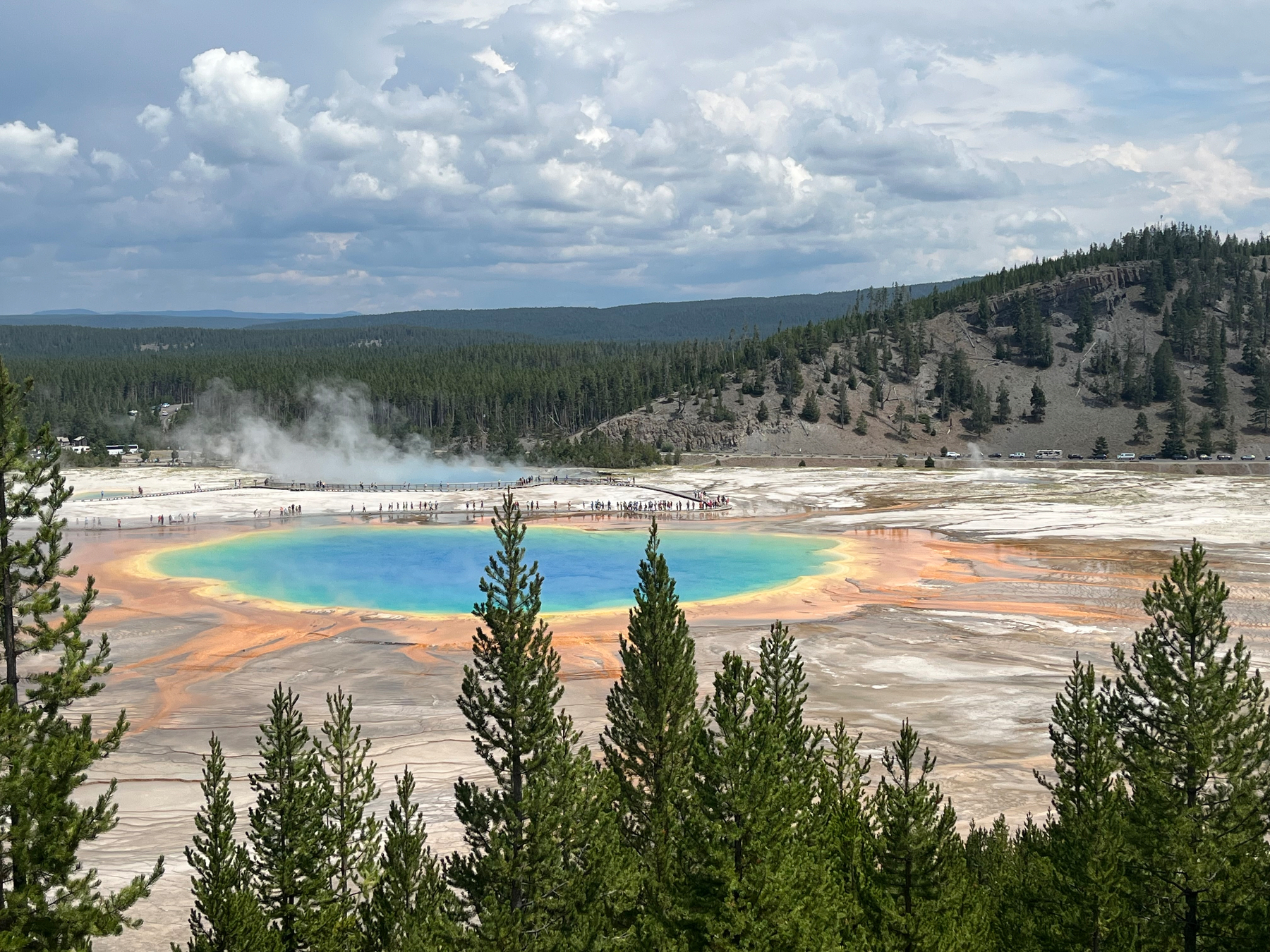 A vibrant, colorful hot spring surrounded by steam, forest, and distant hills is visible.