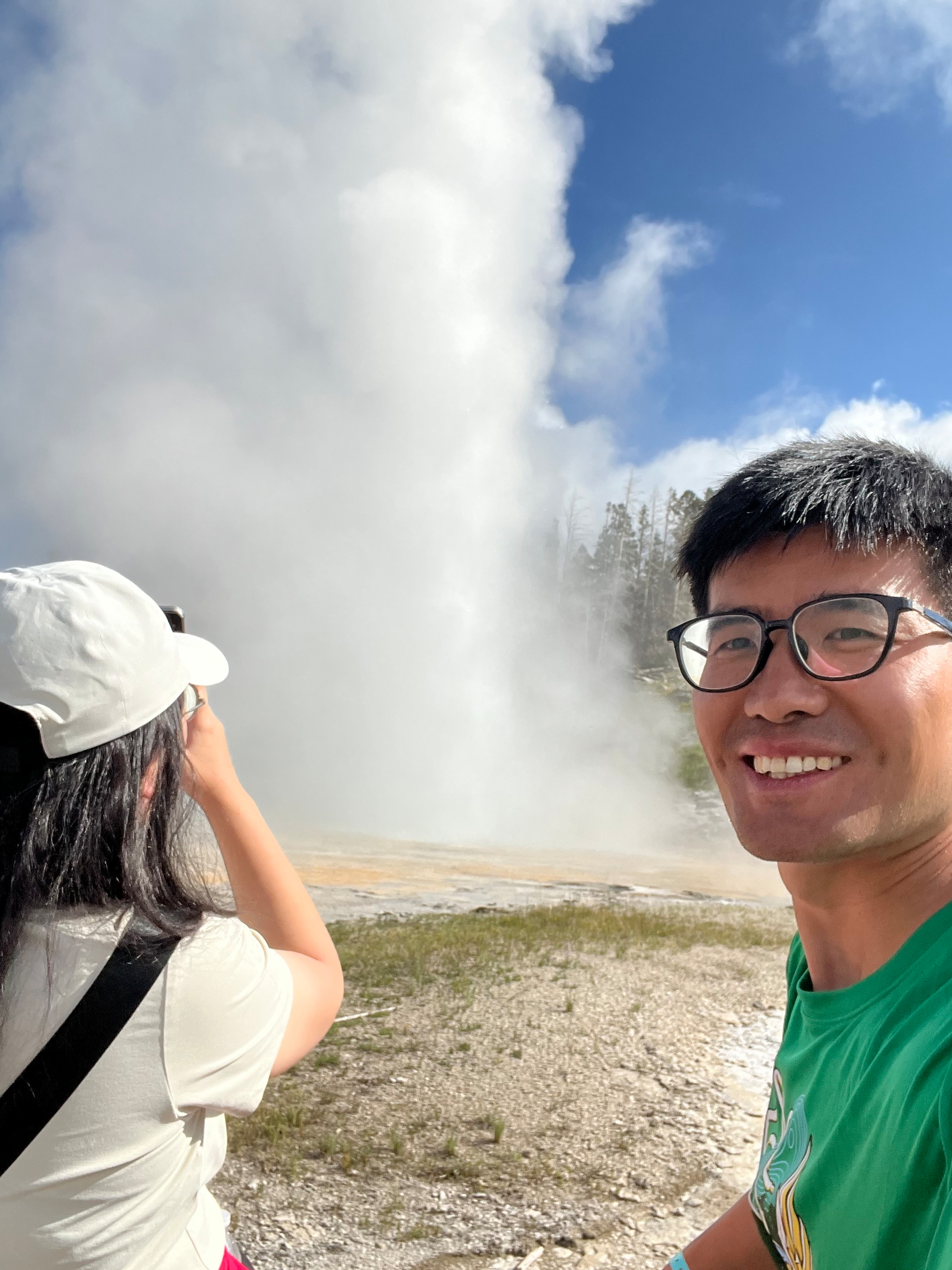 Two people are enjoying a sunny day while capturing photos of an erupting geyser surrounded by a natural landscape.