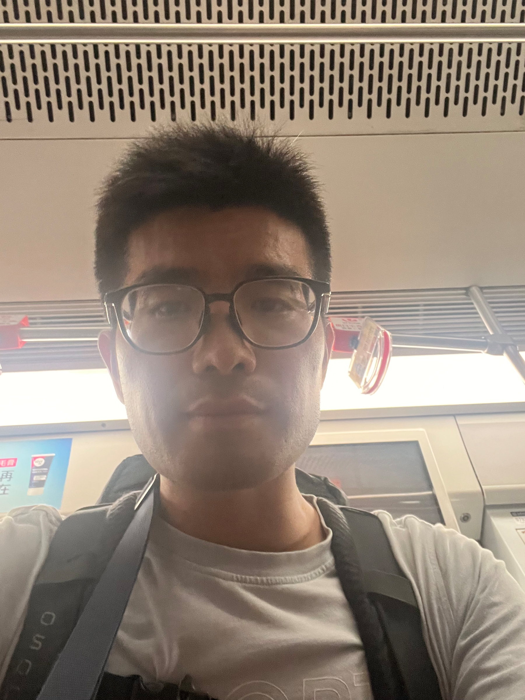 A man wearing glasses, a white T-shirt, and a backpack is taking a selfie inside a public transportation vehicle.