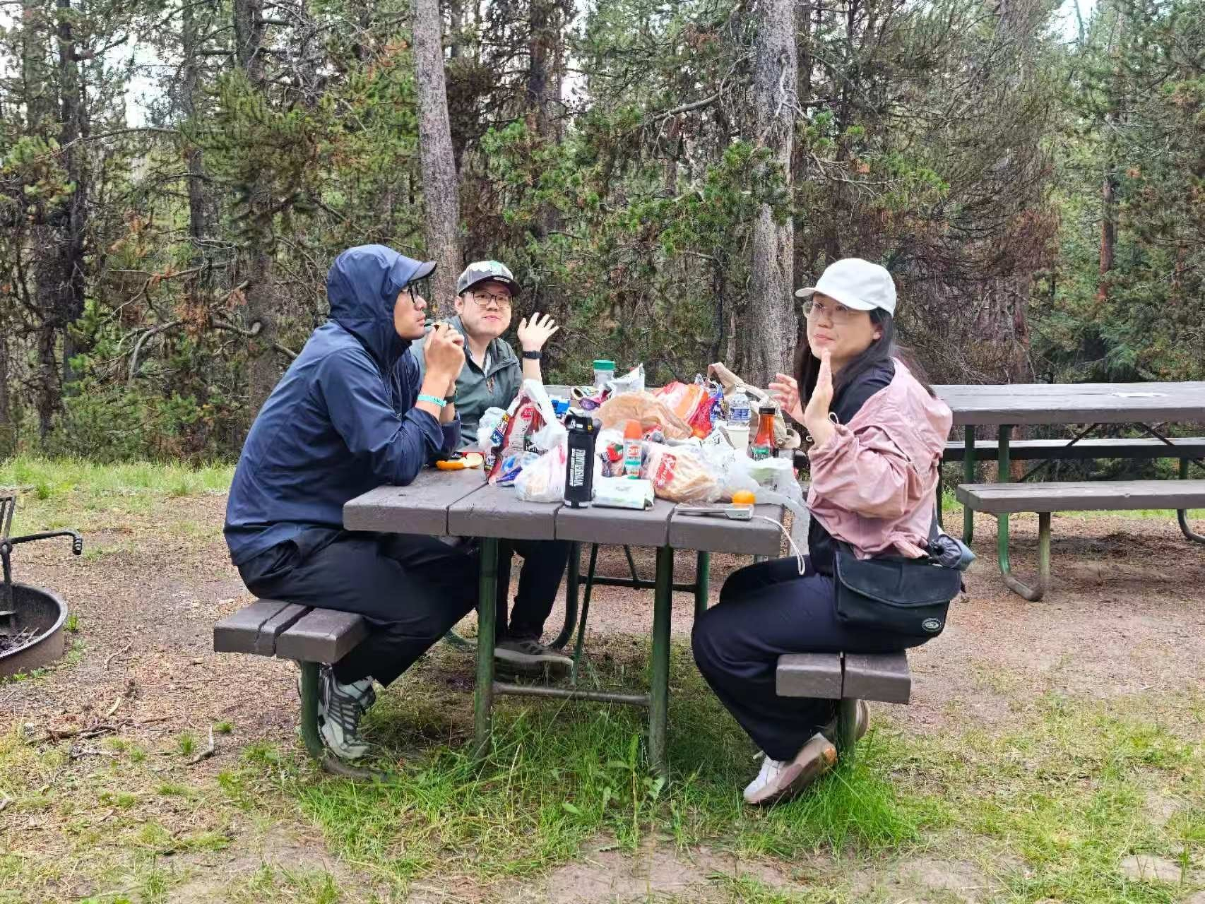 Three people are sitting at a picnic table outdoors, surrounded by trees, enjoying food and drinks.