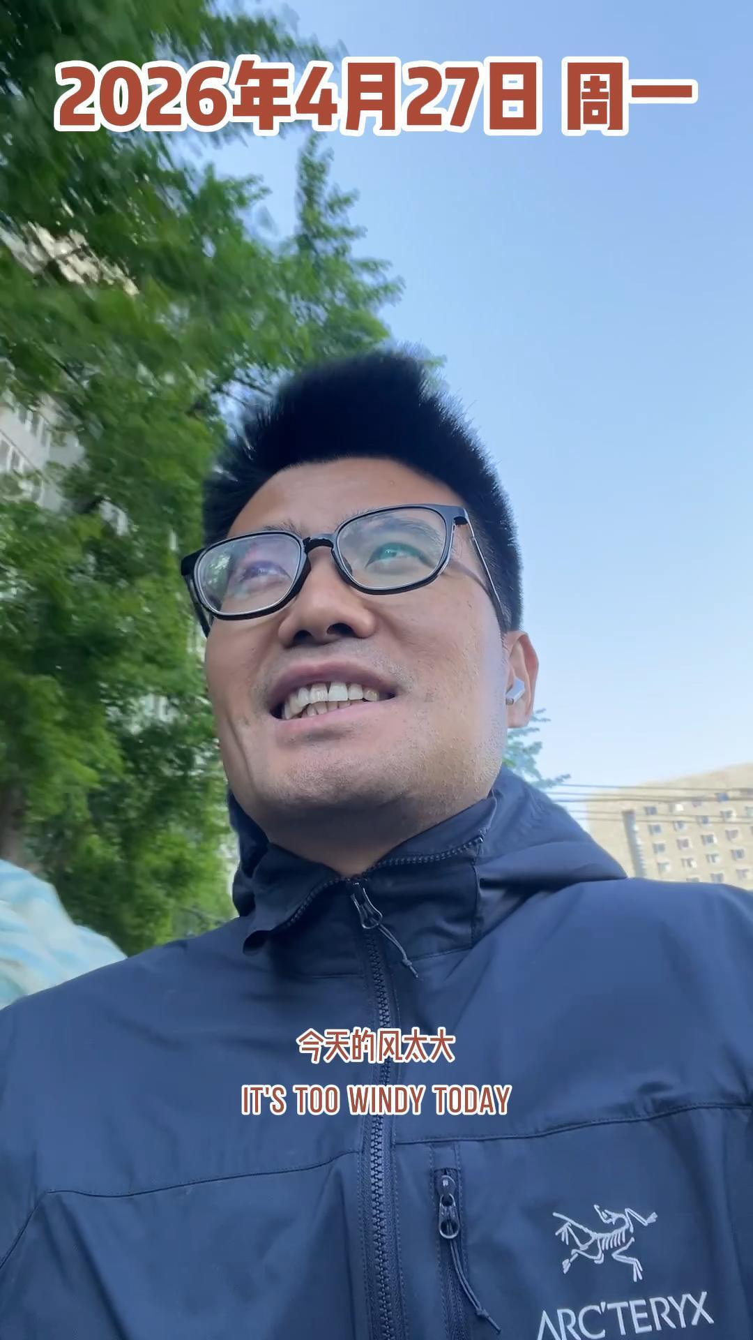 A person wearing glasses and a dark jacket is outdoors on a windy day, with text indicating the date and a comment about the wind.