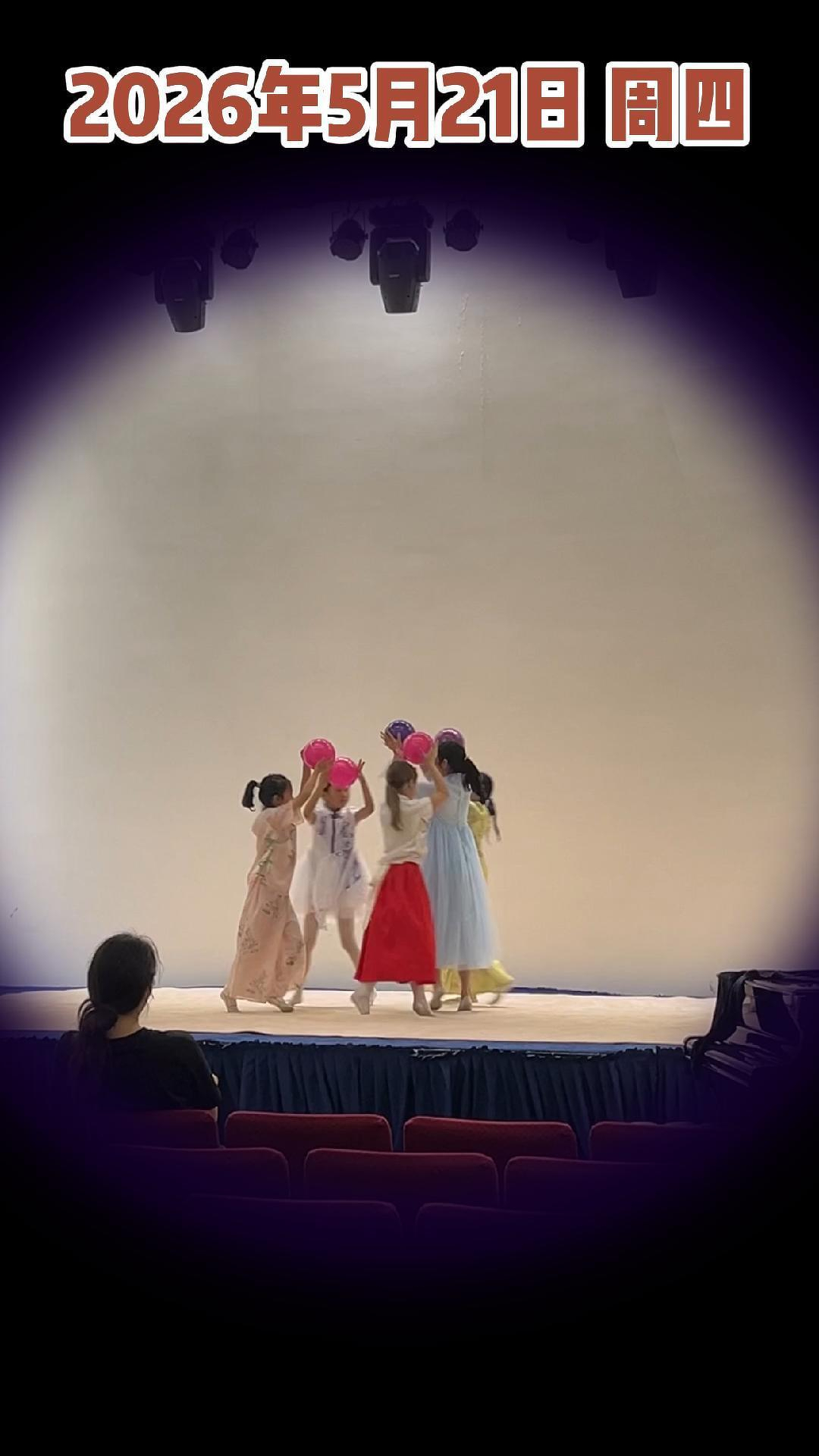 A group of children in colorful costumes are performing on stage under bright lights, with an audience member seated in the foreground.