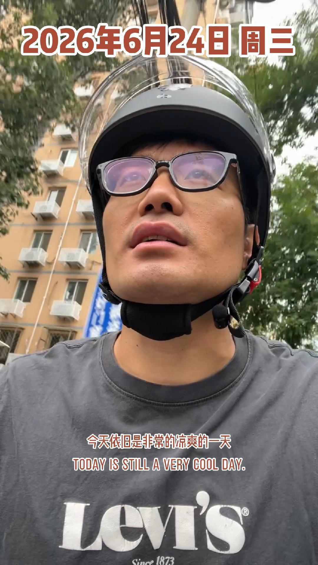 A man wearing glasses and a helmet is outdoors, with buildings and trees in the background, accompanied by text indicating the date as June 24, 2026, and stating that it is still a very cool day.