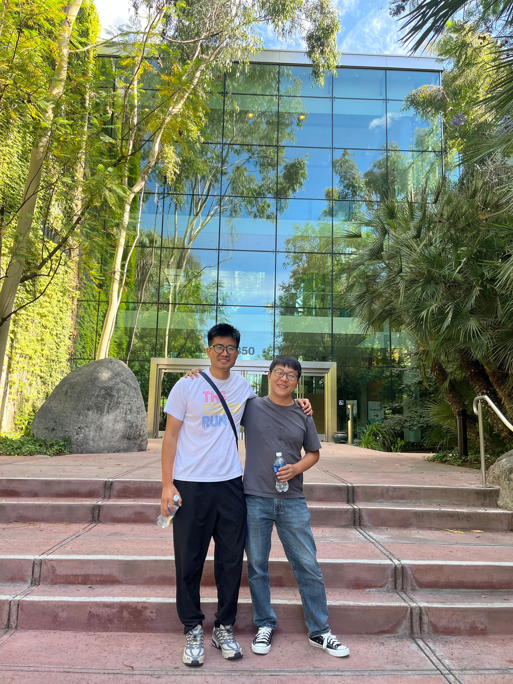 Two young men smiling and posing together on steps in front of a modern glass building surrounded by greenery.