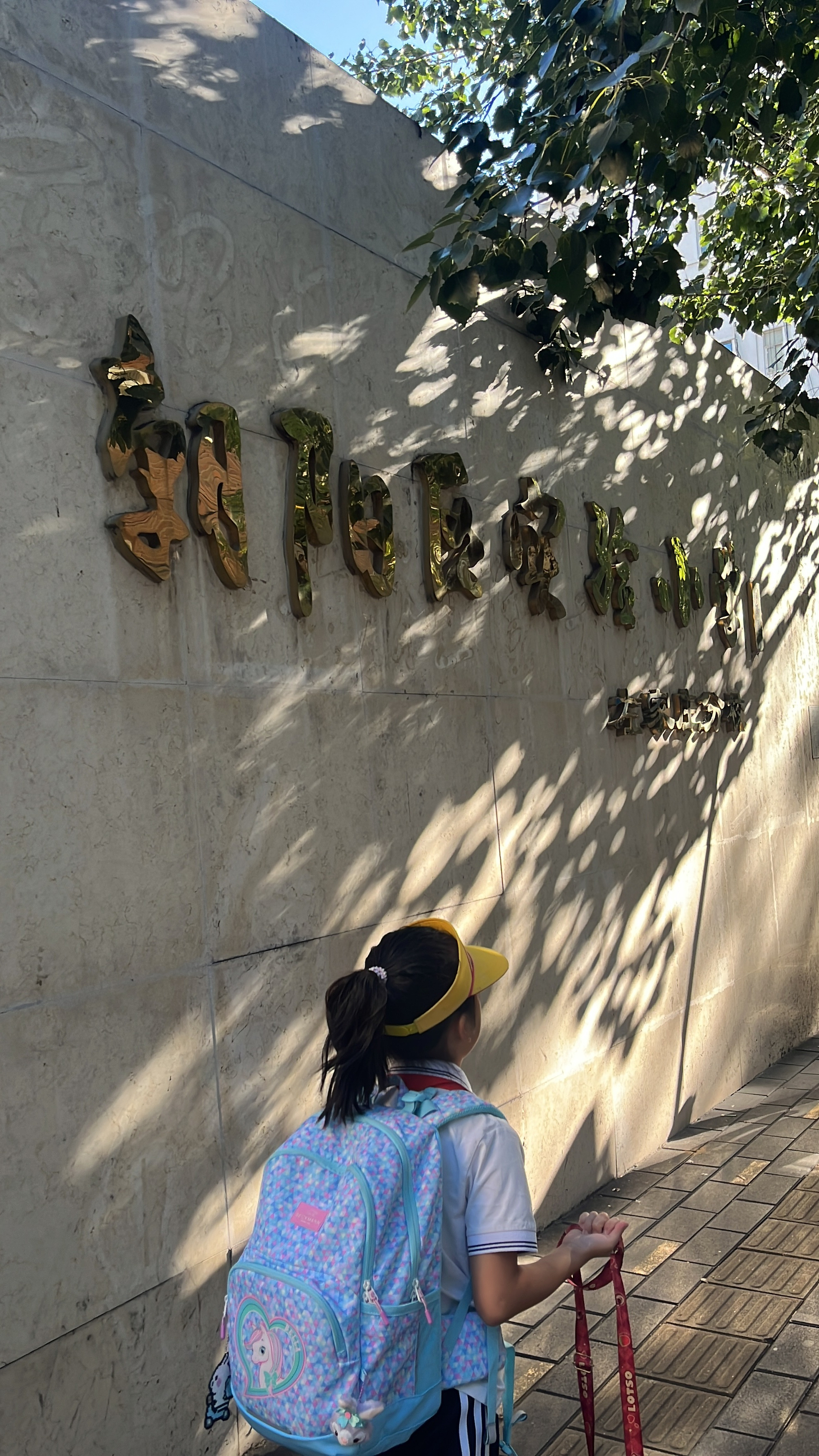 A child wearing a yellow visor and carrying a colorful backpack stands under the shade of trees next to a wall with gold-colored Chinese characters.