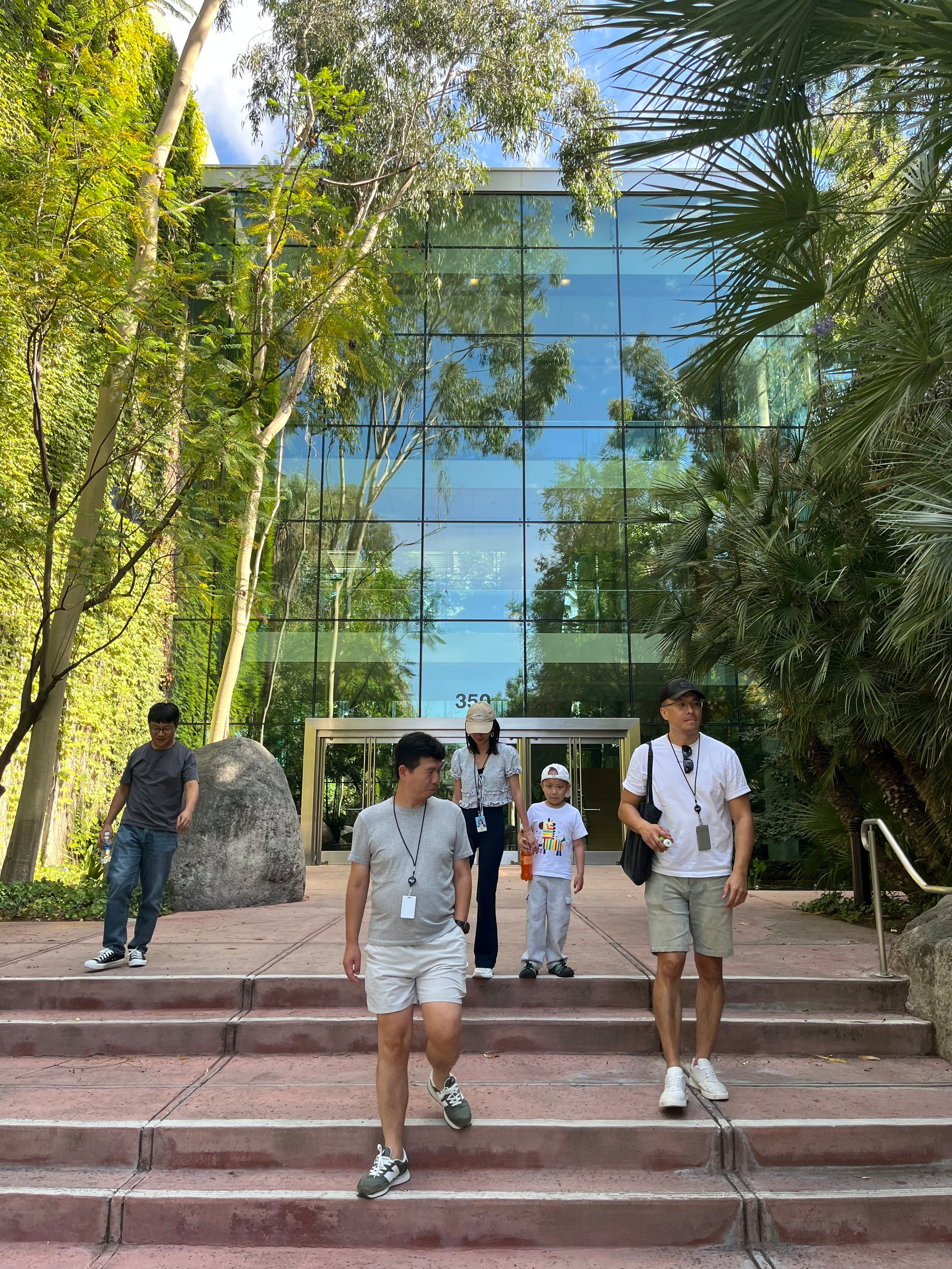 Five people are walking down the steps in front of a modern building with reflective glass windows surrounded by lush greenery.