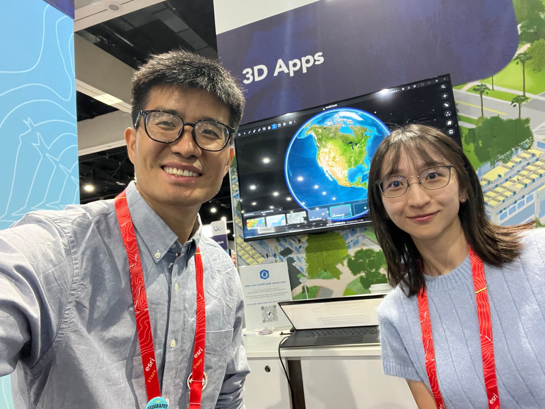 Two people wearing glasses and red lanyards are smiling in front of a display screen showing a 3D map of North America labeled 3D Apps.