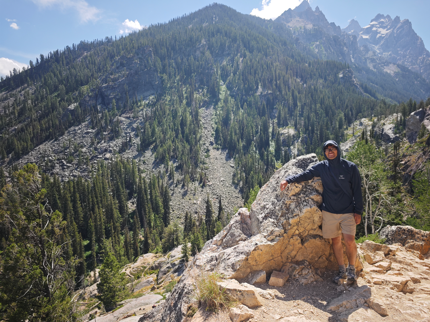 A person dressed in outdoor clothing stands on a rocky trail overlooking a forested mountainous landscape under a partly cloudy sky.
