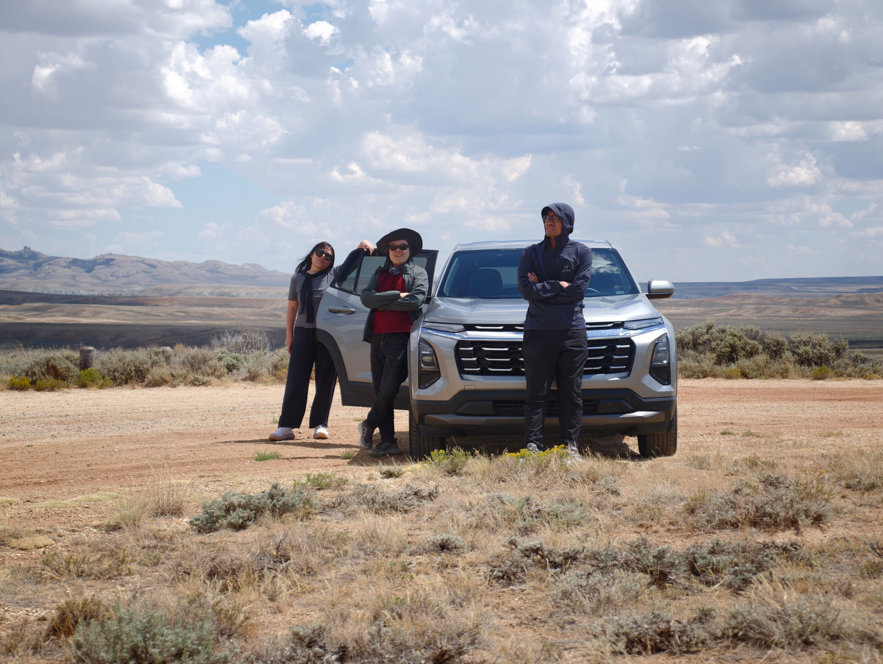Three people stand around and in front of a silver SUV in a vast, open desert landscape under a partly cloudy sky.