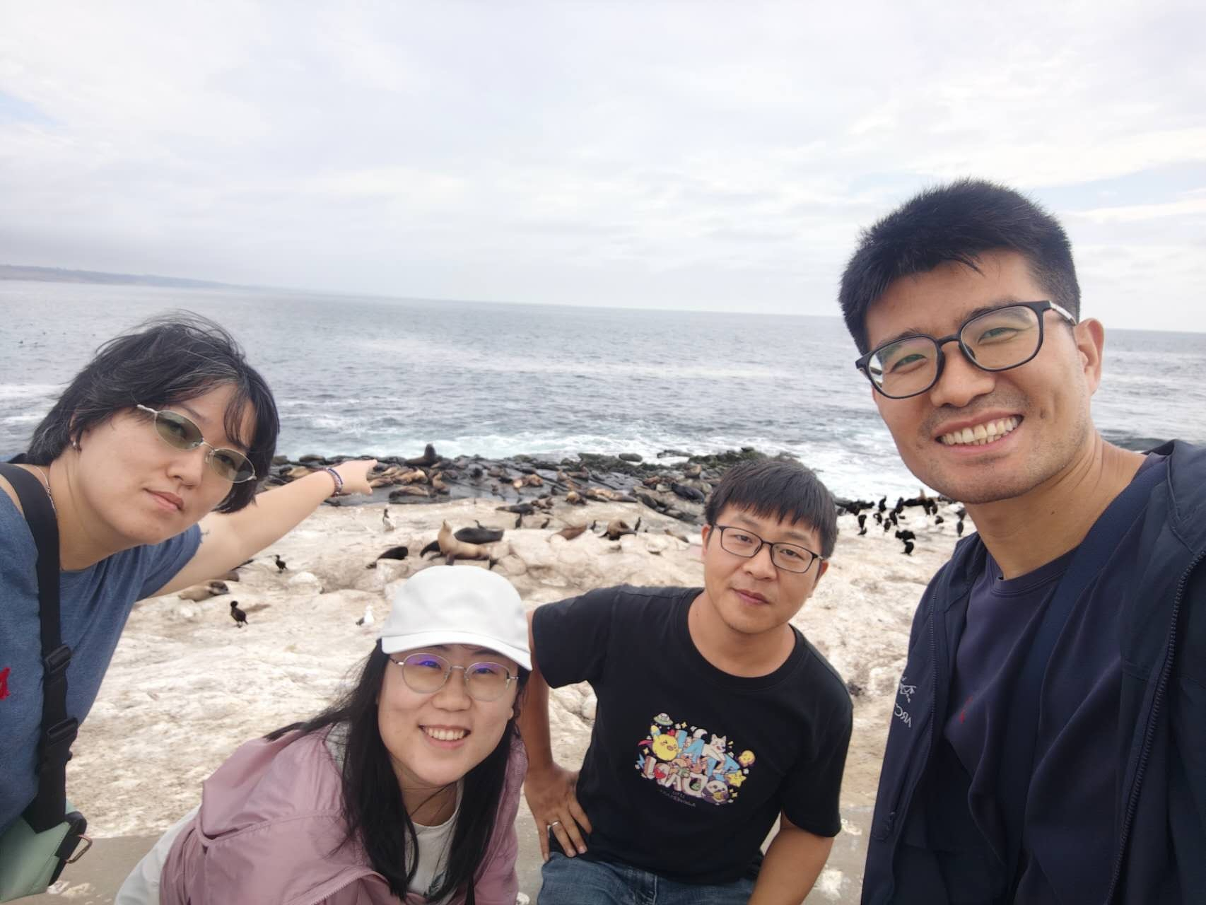 Four people are posing for a selfie on a rocky shoreline with seals and birds resting in the background near the ocean.
