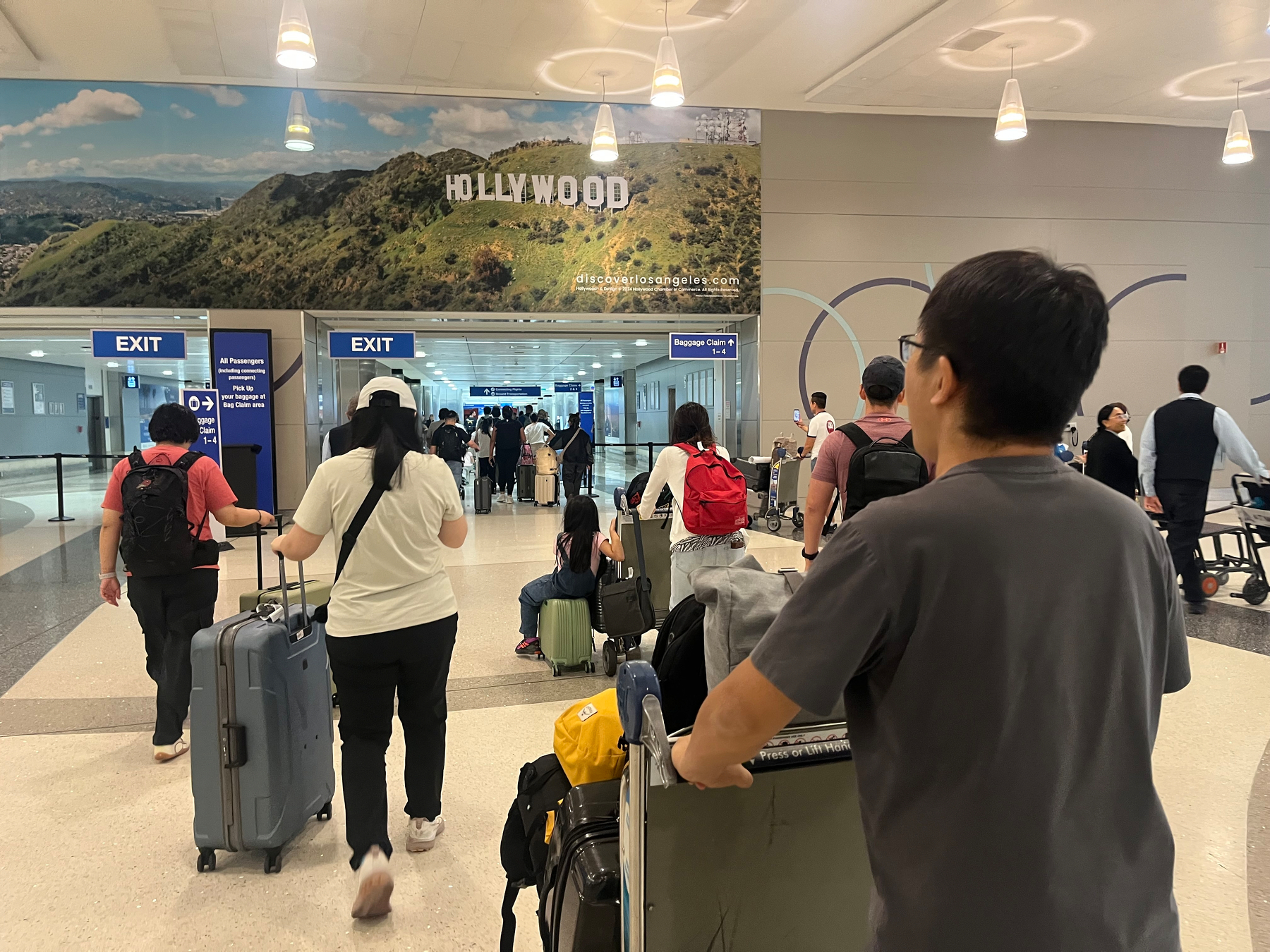 People with luggage and backpacks are walking towards the exit area inside an airport terminal, with a large Hollywood sign poster displayed overhead.
