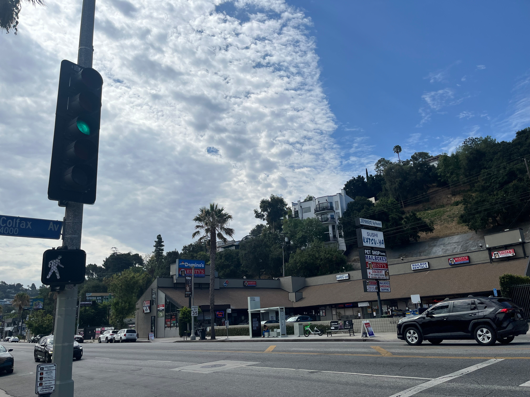 A street intersection with a green traffic light, a pedestrian signal, a shopping center in the background, and a partly cloudy sky.