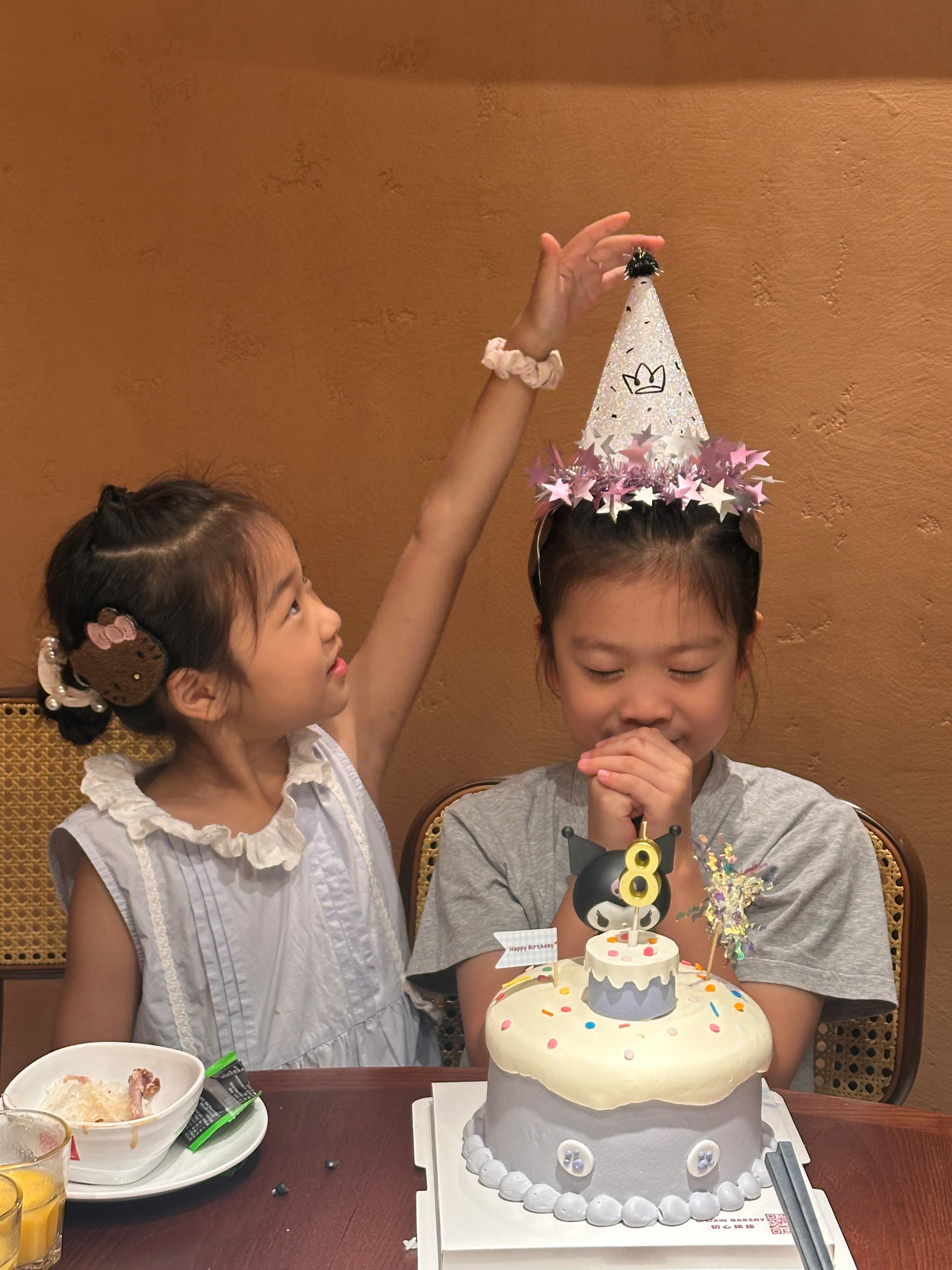 Two young girls are celebrating a birthday, with one wearing a party hat and the other reaching to adjust it while a cake with a number 8 candle is on the table.
