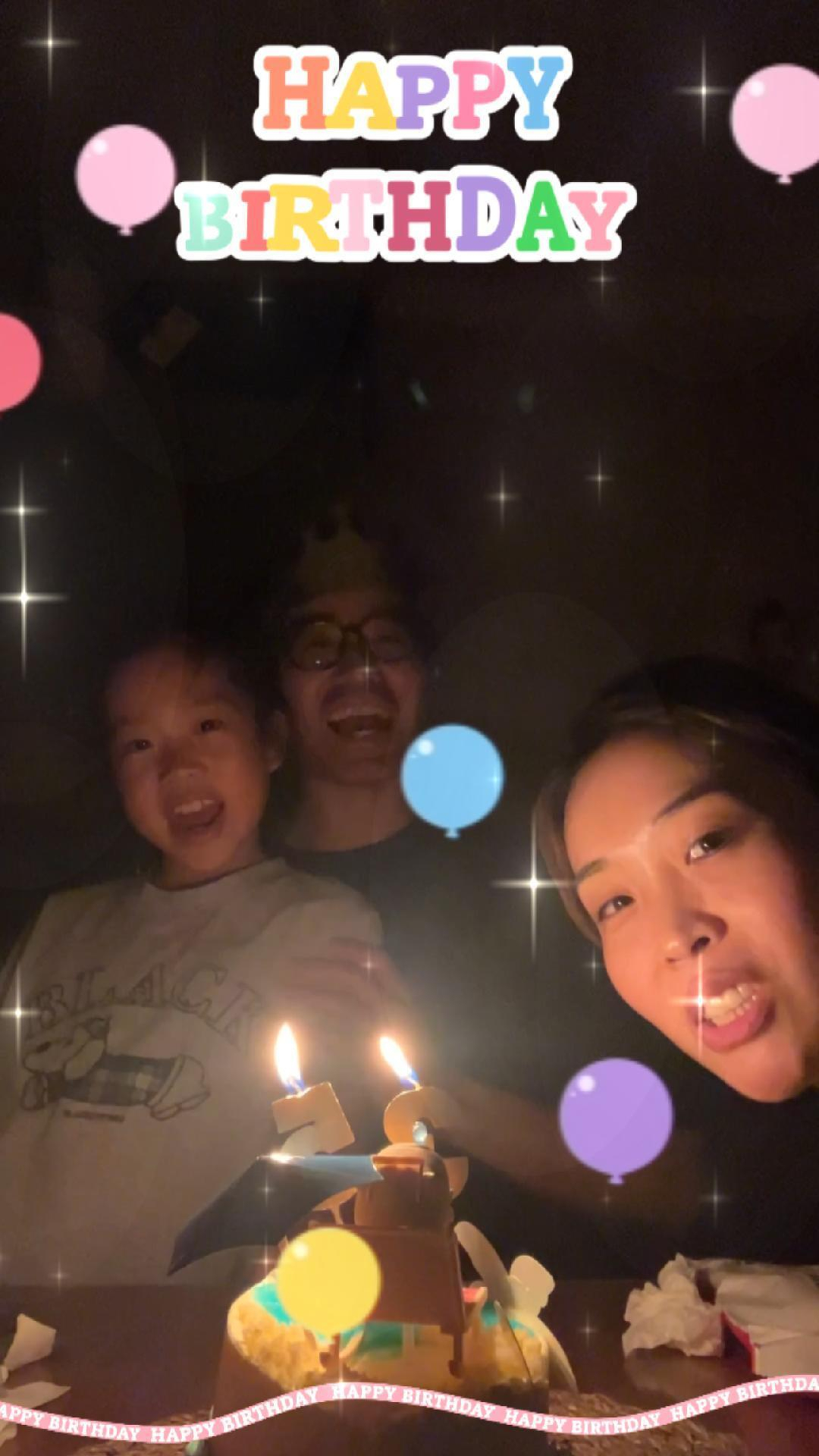 A family is gathered around a lit birthday cake, smiling and celebrating together.