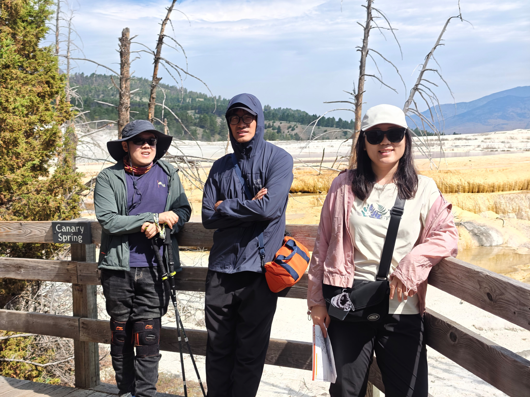 Three people dressed in outdoor clothing and hats are standing on a wooden platform near a sign that says Canary Spring with a natural landscape of trees and hills in the background.