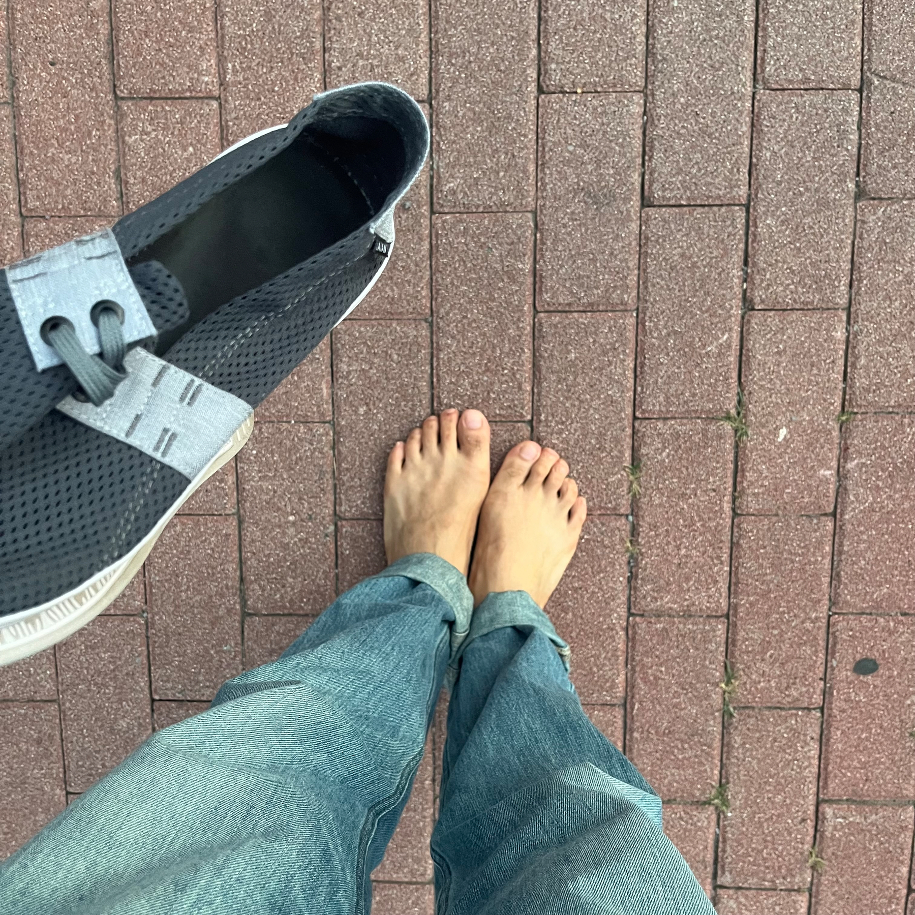 A person is standing barefoot on a brick pavement while holding a gray shoe in one hand.