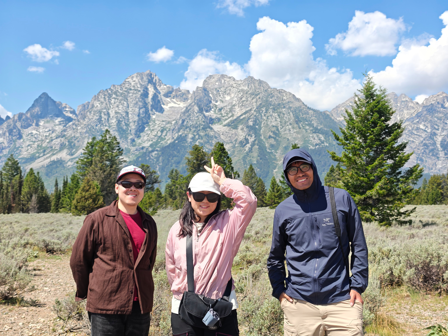 Three people are posing and smiling in front of a scenic mountain range under a partly cloudy sky.