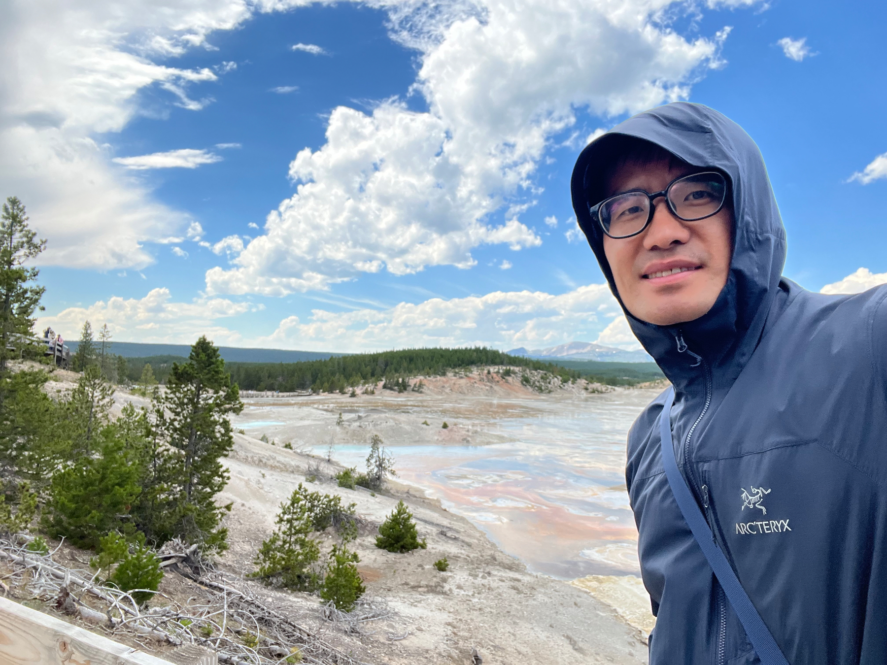 A person wearing glasses and a hooded jacket stands outdoors near a geothermal area with trees and a partly cloudy sky in the background.
