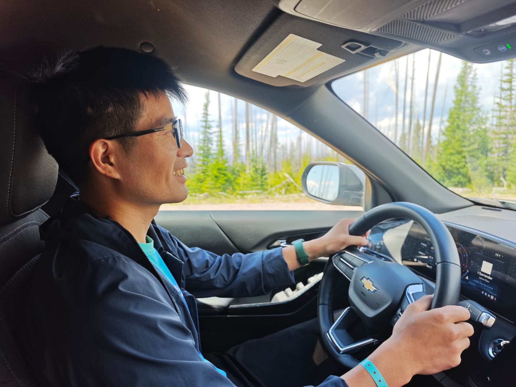 A person wearing glasses and a dark jacket is smiling while driving a Chevrolet car through a forested area.