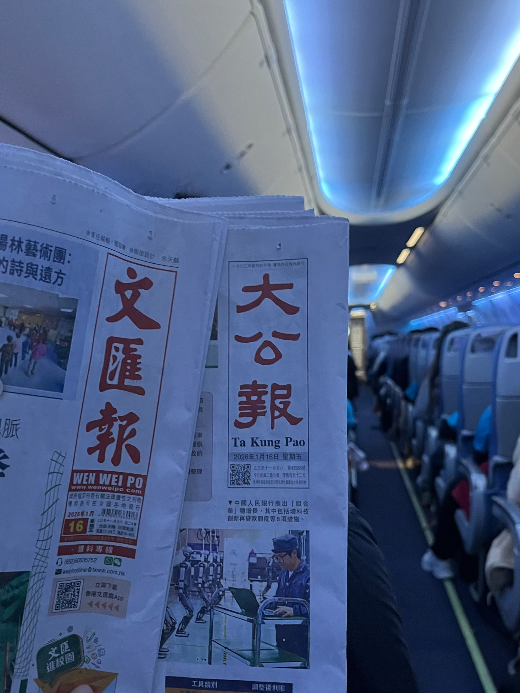 A person is reading a newspaper inside an airplane cabin with blue ambient lighting.