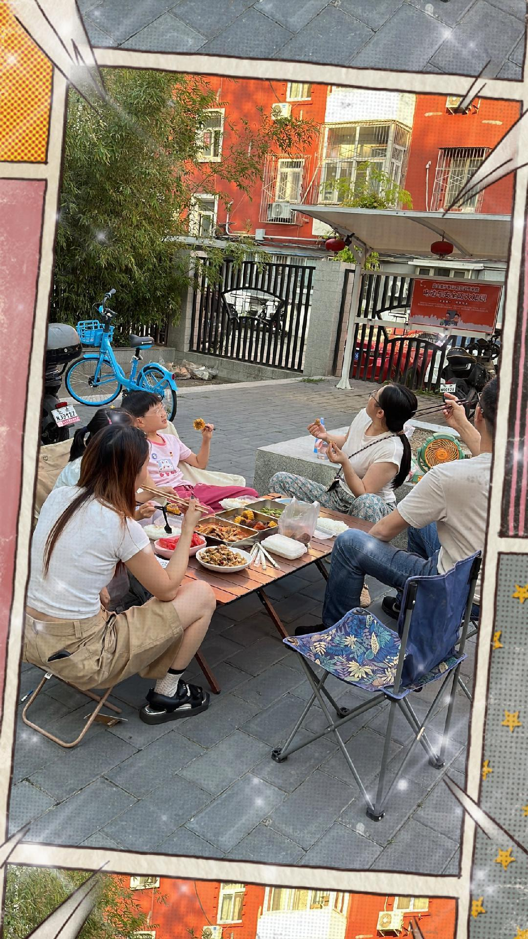 A group of people is enjoying a meal together outdoors while sitting on foldable chairs around a low table.