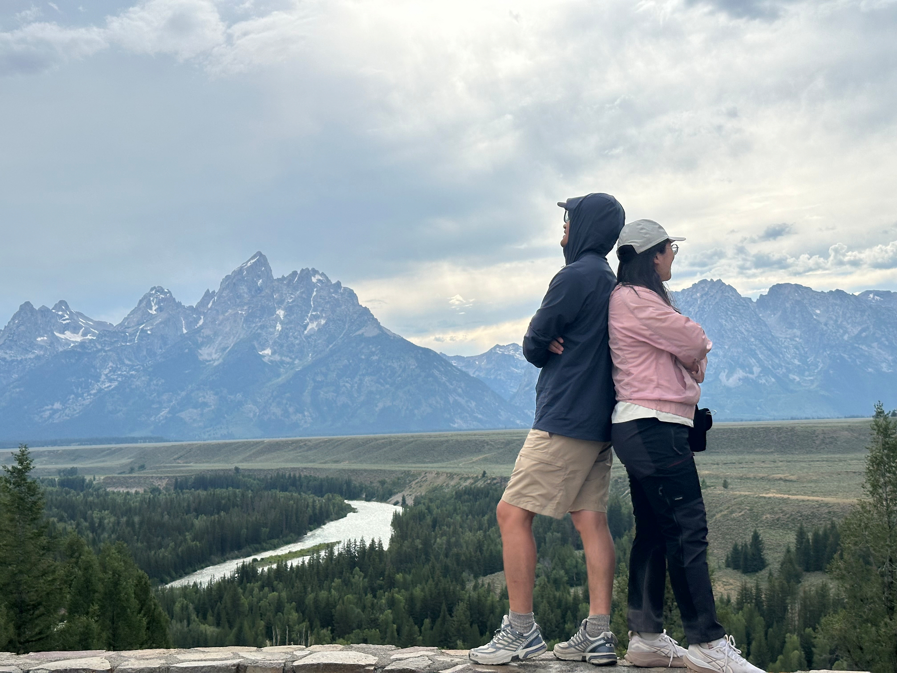 Two people stand back-to-back on a scenic overlook with a river and mountain range in the background under a cloudy sky.
