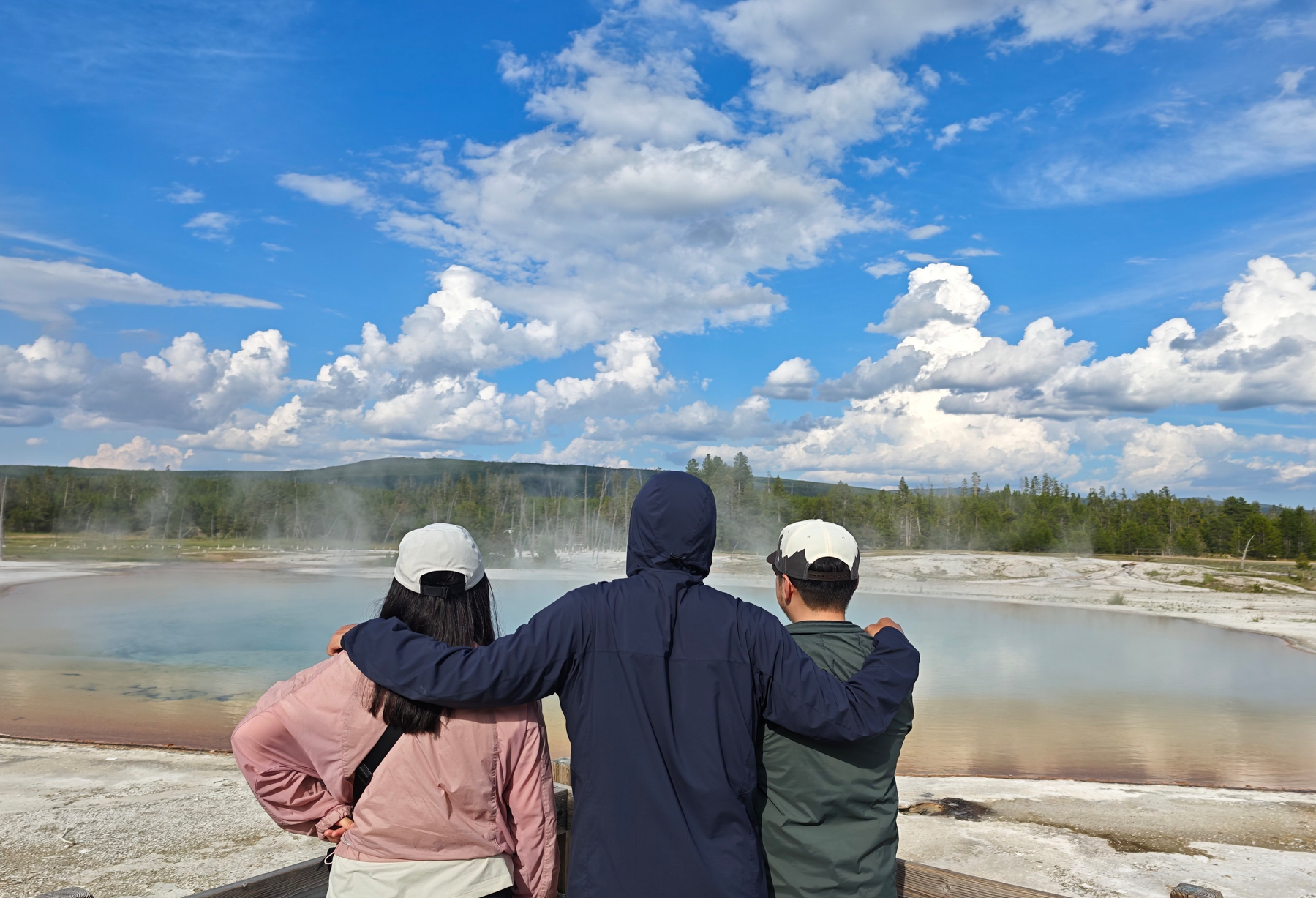 Three people stand together with their arms around each other, looking out at a steaming geothermal pool under a partly cloudy sky.