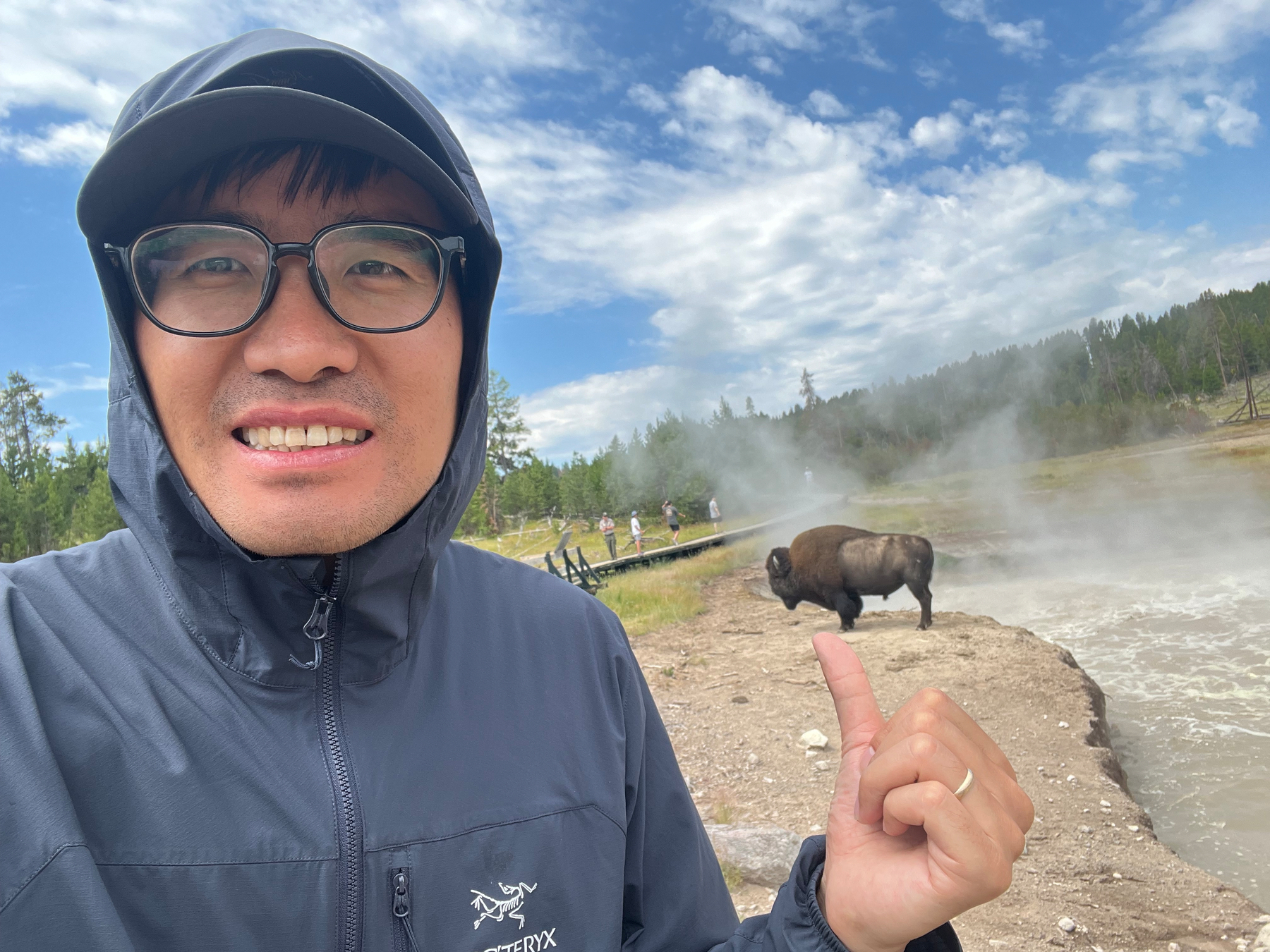 A person wearing glasses and a hooded jacket is posing for a selfie while appearing to hold a bison on their finger in a natural park setting.