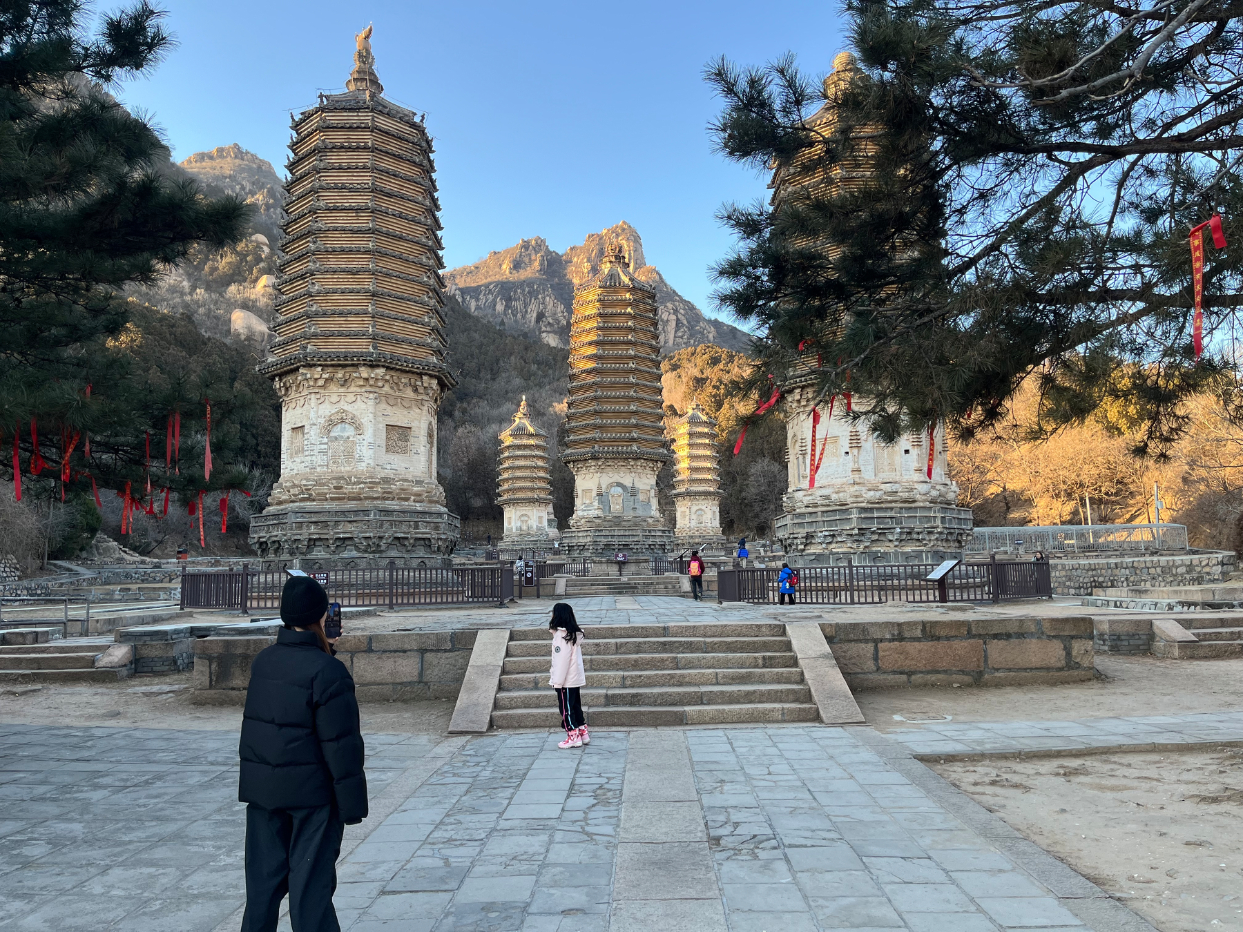 A person in a black jacket and a child stand before ancient pagodas surrounded by trees and mountains, with red ribbons hanging nearby.