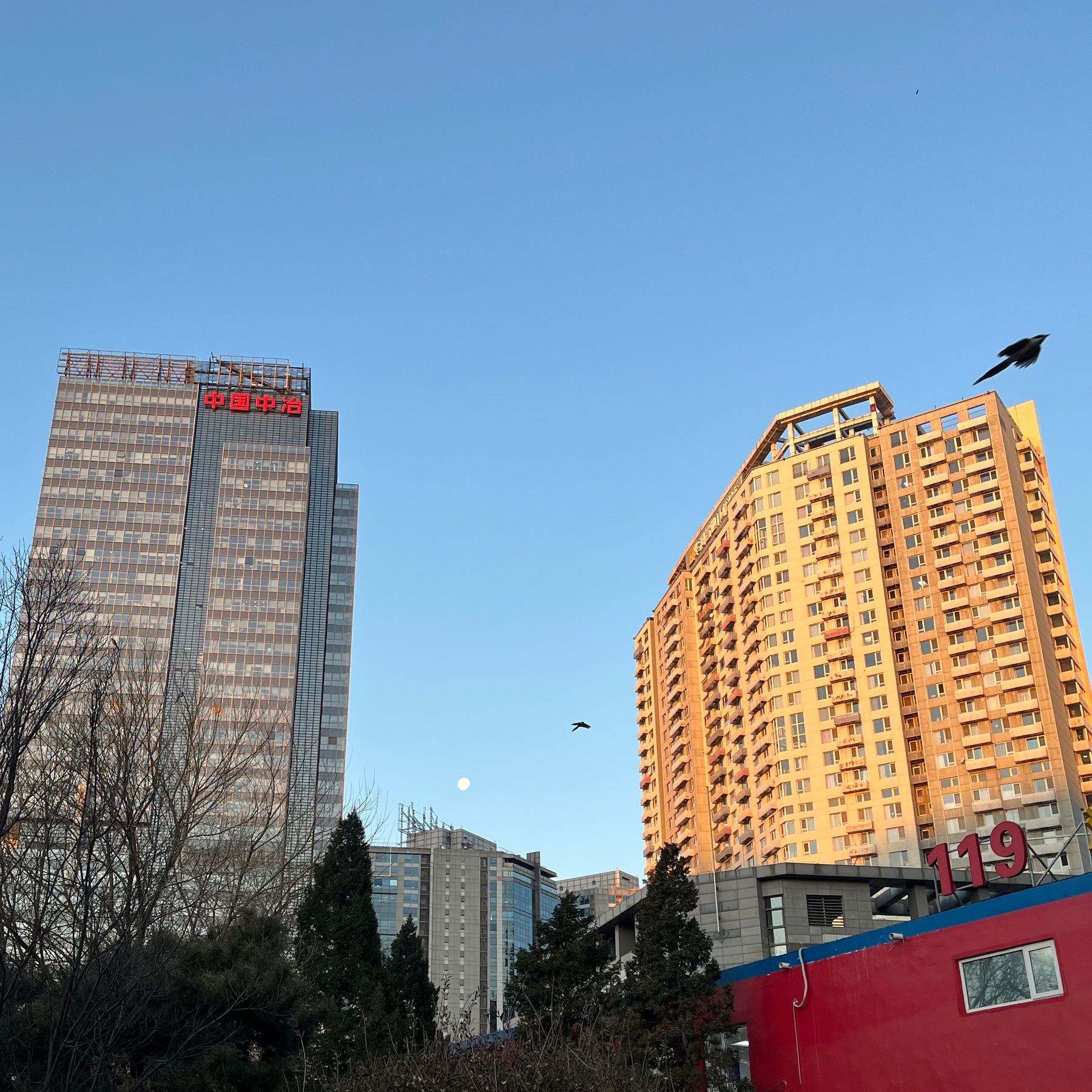 Two tall buildings rise above a cityscape with a clear sky, while a red structure and some trees are visible in the foreground.