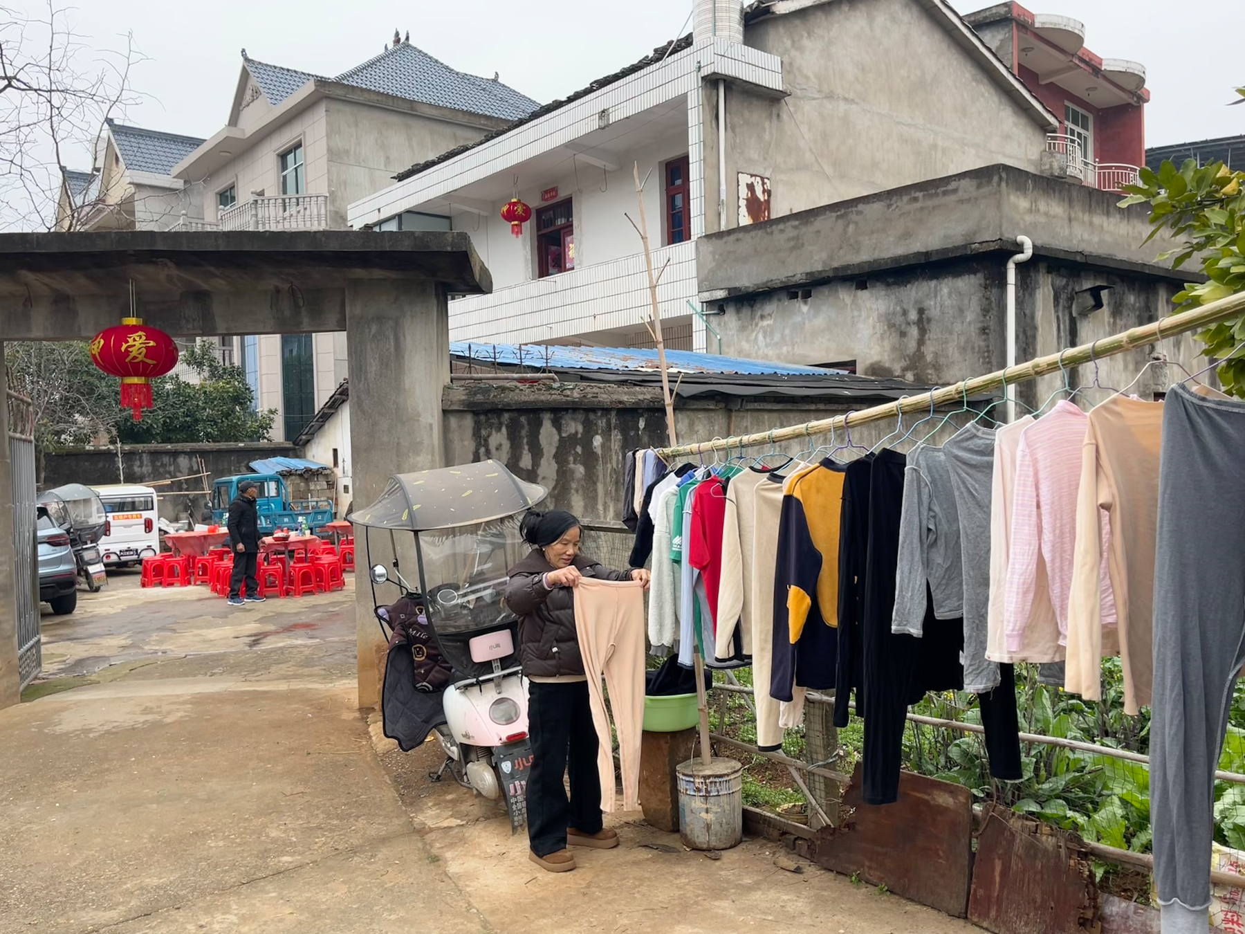 A person is hanging clothes on a line in a residential area with houses and vehicles in the background.