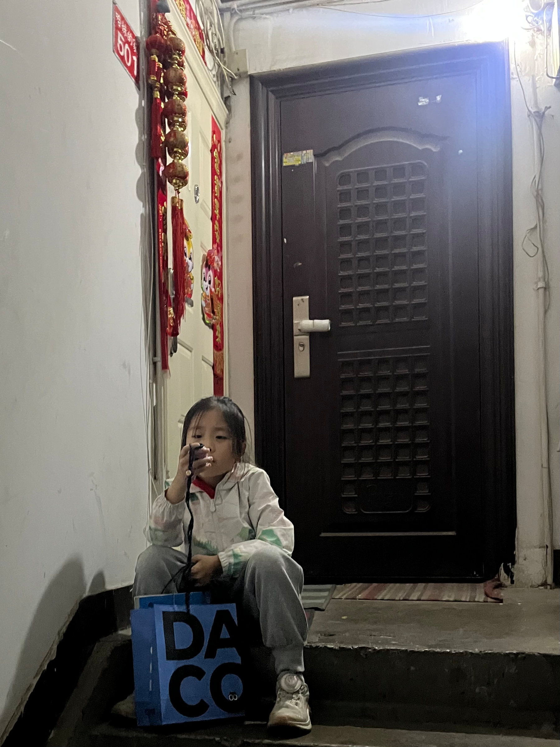 A child sits on a staircase holding a phone with a blue bag labeled DACO beside them.