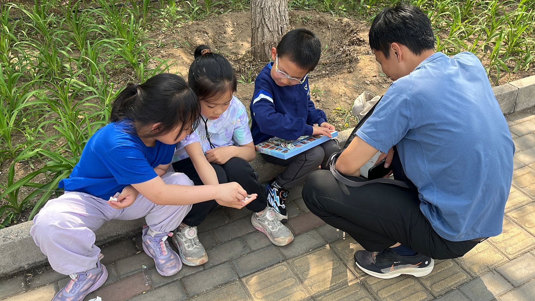A man kneels on a sidewalk, engaging with three children who are seated and looking at a picture book.