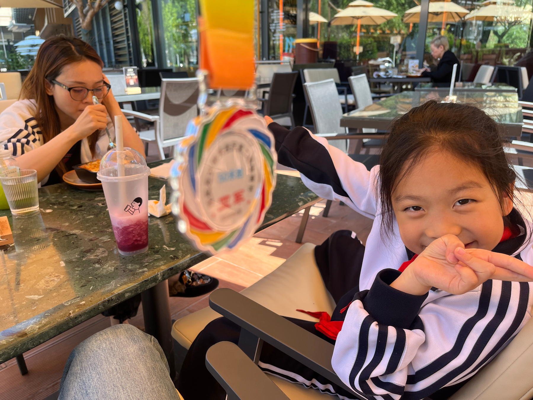 A young girl smiles playfully at the camera while sitting at an outdoor restaurant, with another person eating beside her and a colorful medal hanging in the foreground.