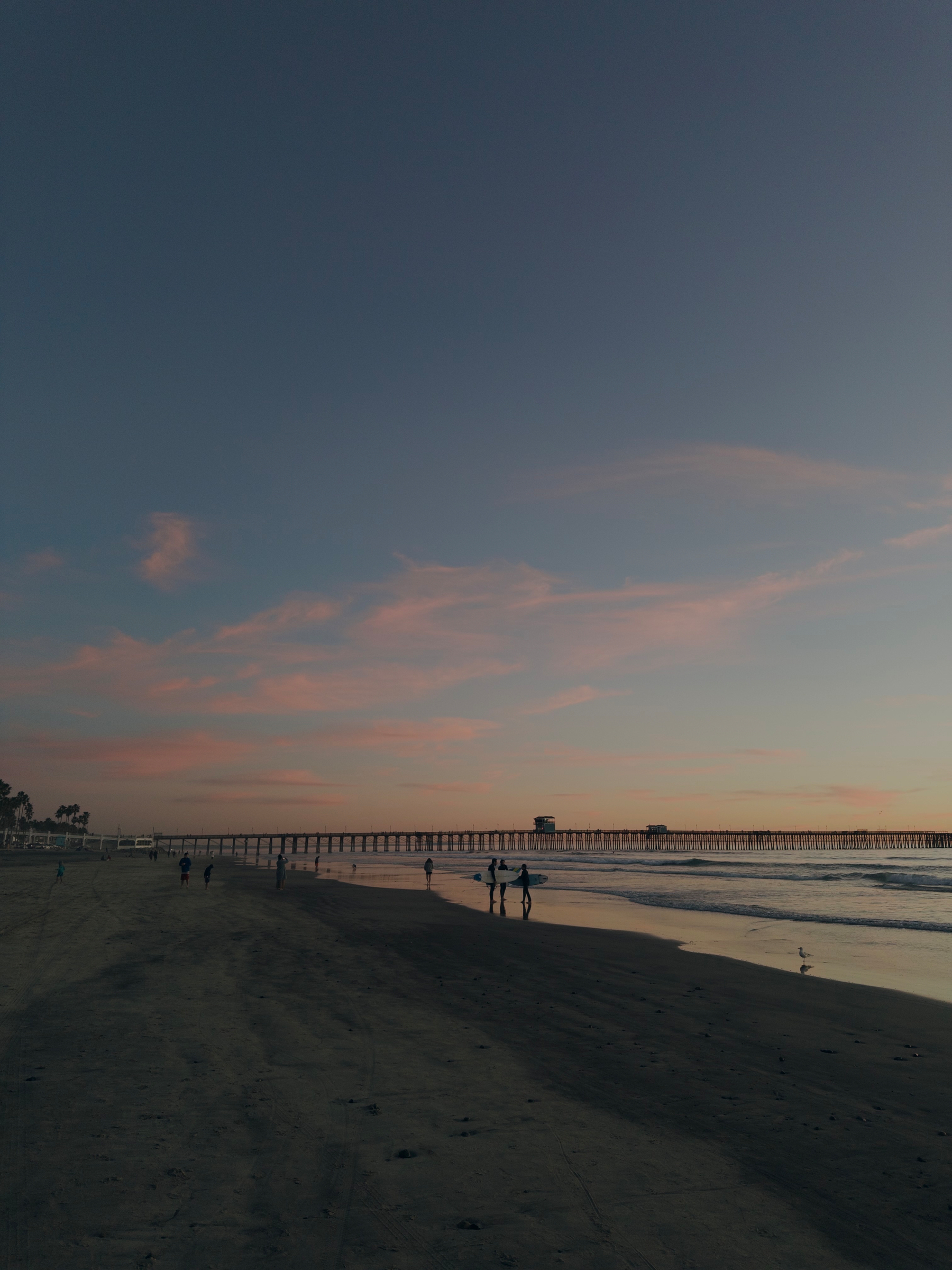 A tranquil beach scene at sunset features silhouettes of people walking along the sand and a long pier extending into the ocean.