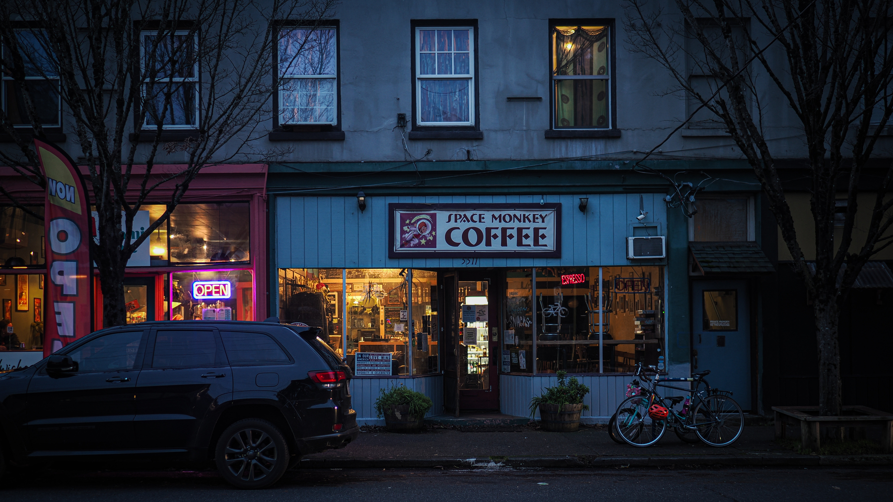 A cozy coffee shop named Space Monkey Coffee is warmly lit from the inside, with a parked car and bicycles outside in an urban evening setting.