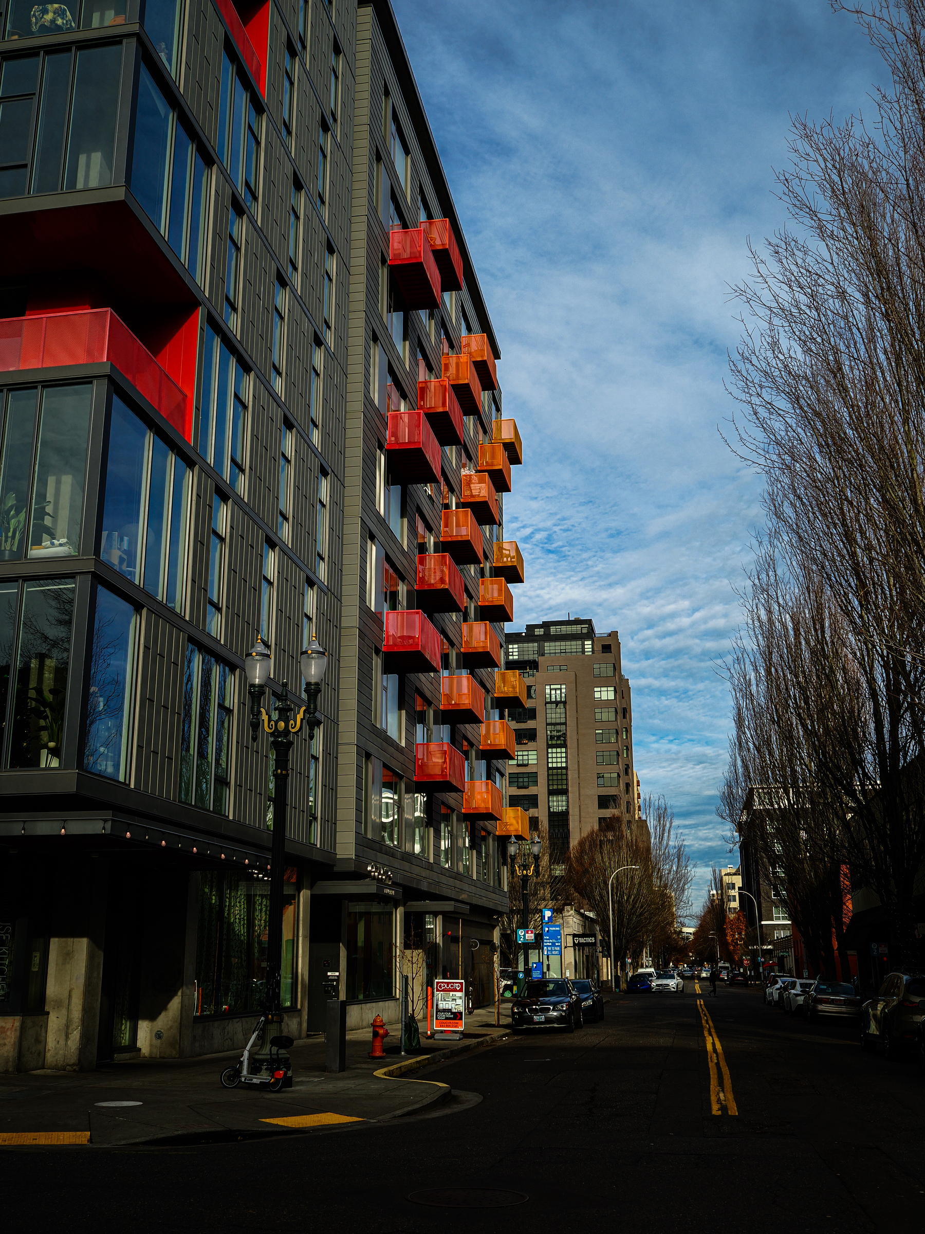 A city street features a modern building with bright red balconies under a partly cloudy sky.