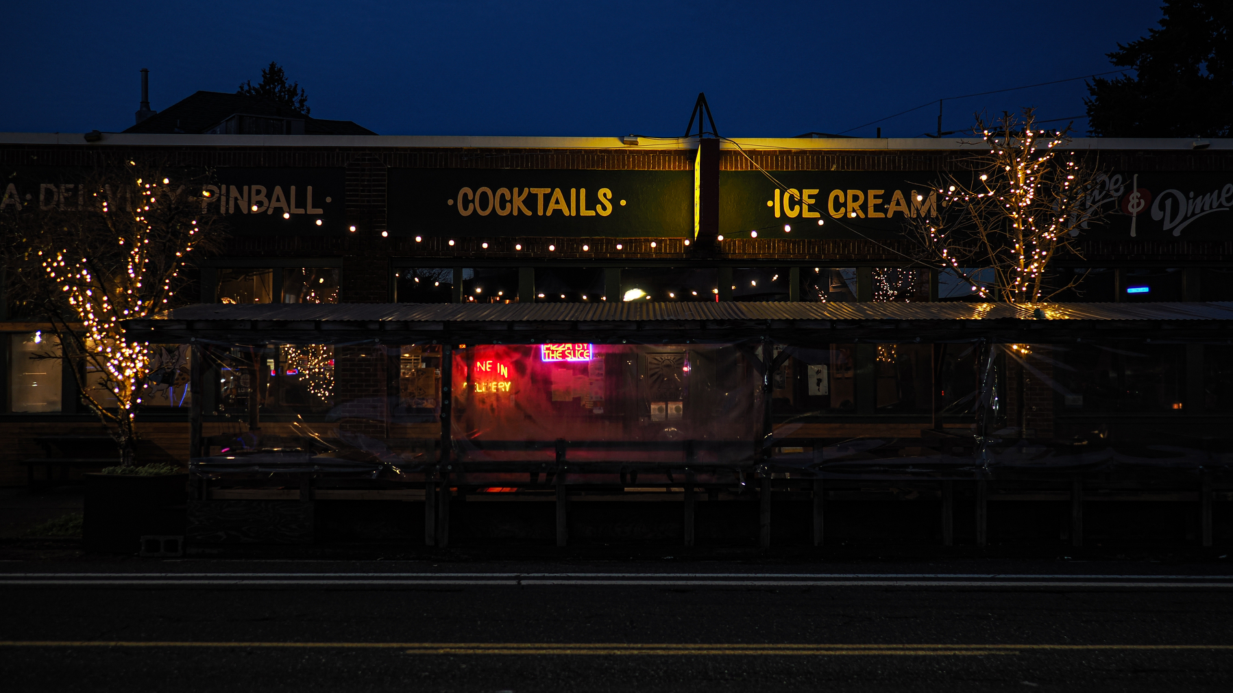 A warmly lit storefront featuring signs for pinball, cocktails, and ice cream is adorned with string lights and neon signs at night.