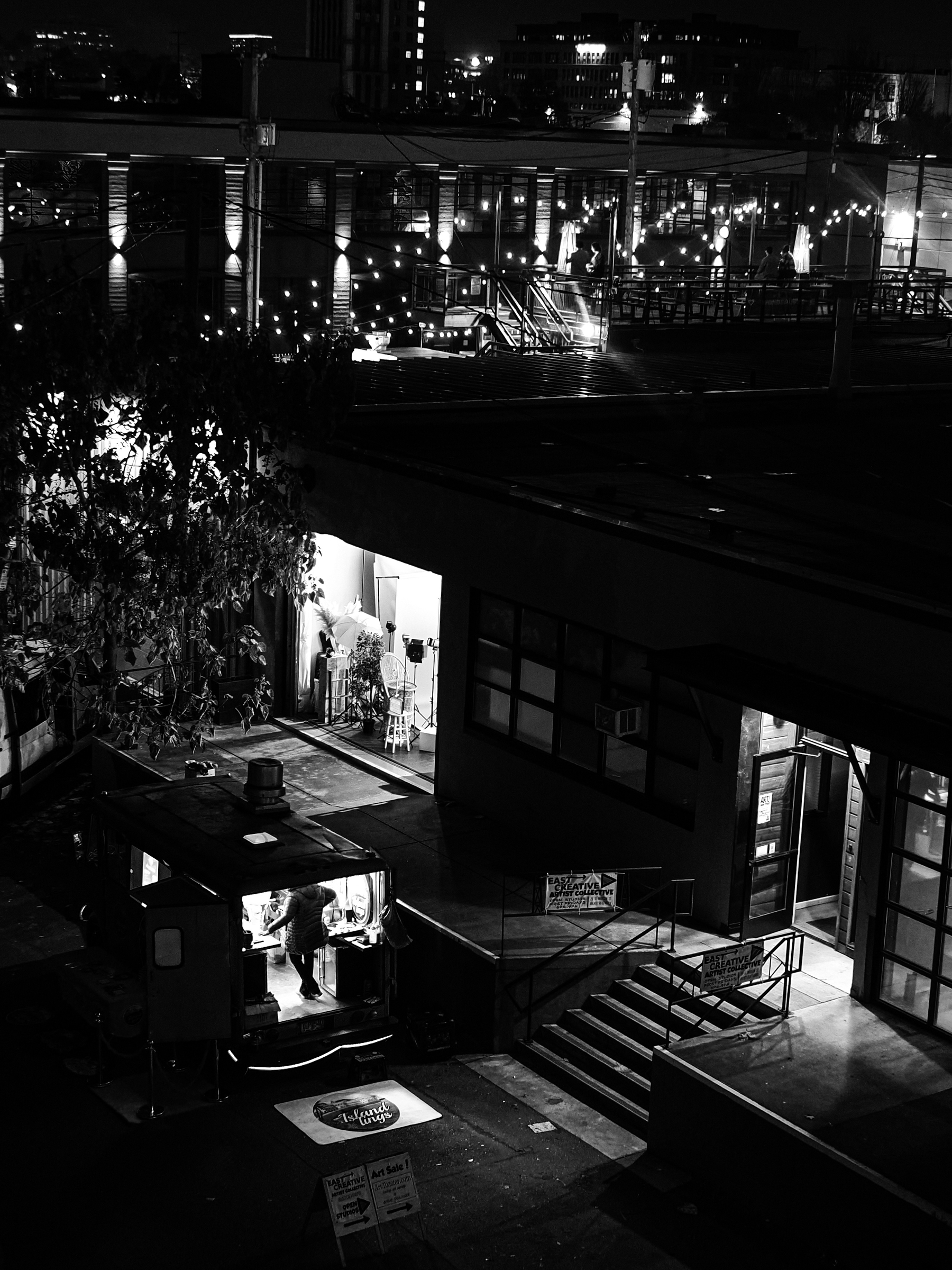 A nighttime scene in a city features a small food truck with a person inside, surrounded by dimly lit buildings and string lights.