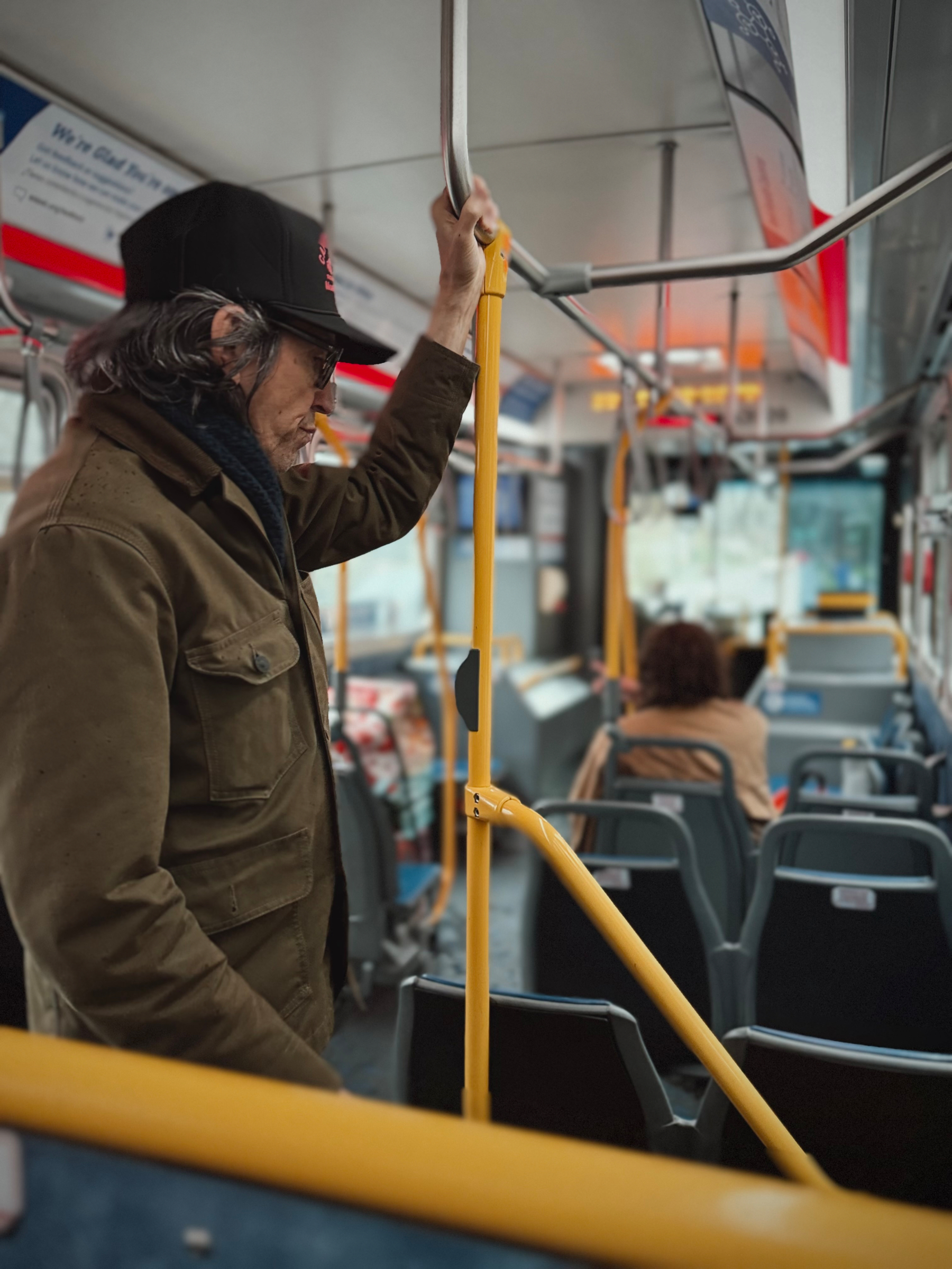 A person wearing a hat and jacket stands inside a bus, holding onto the overhead railing, with several seats and another passenger visible in the background.