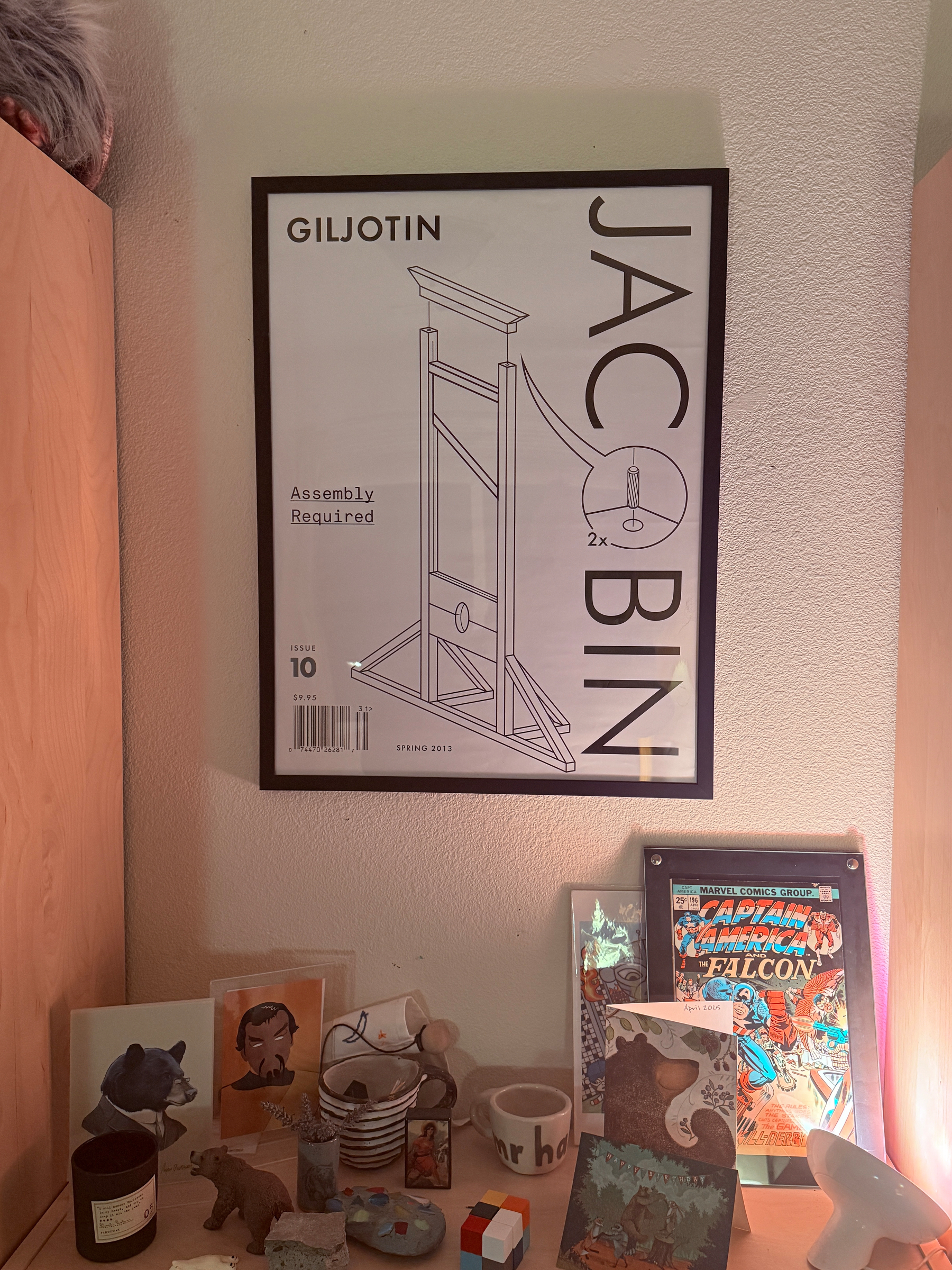 A framed poster featuring a stylized guillotine design titled GILJOTIN - Assembly Required hangs above a cluttered shelf with books, figurines, and artwork.